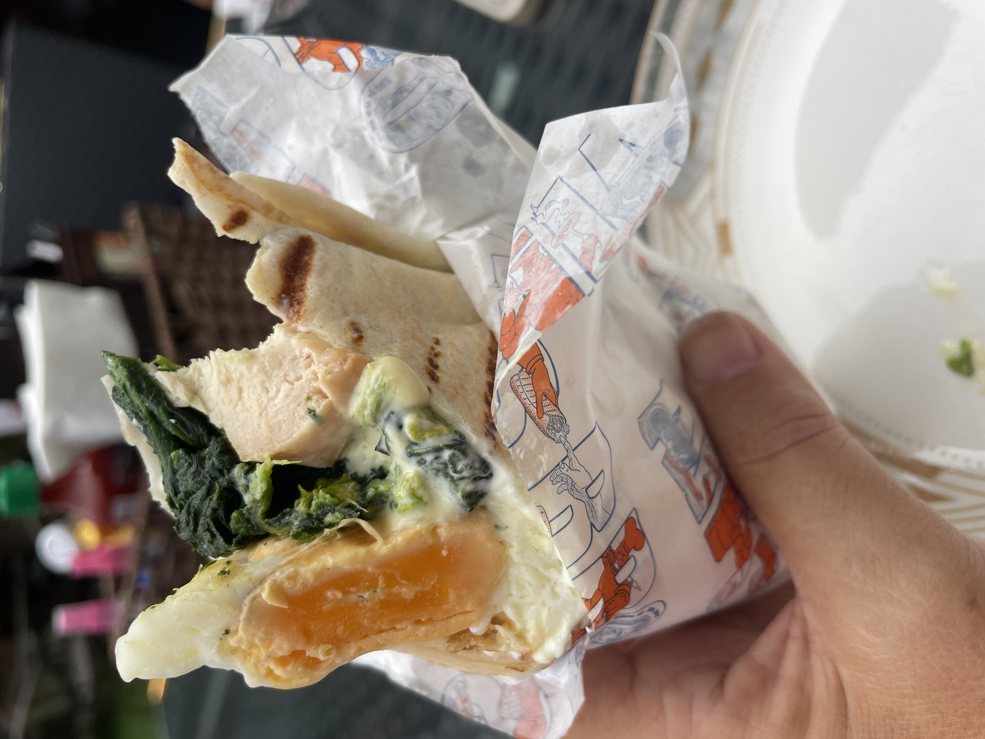 A hand holds a partially unwrapped wrap or burrito-style food item, presumably in Choeng Thale, Thailand, given the prompt. The wrap contains a bright orange egg yolk, cooked spinach, pieces of what appears to be chicken, and a creamy white sauce. The wrap itself is lightly browned. The wrapping paper is white with images of cartoon chefs and lobsters, predominantly in orange and blue. The background is blurred, suggesting an outdoor or semi-outdoor environment, with hints of foliage and other objects that are indistinguishable.