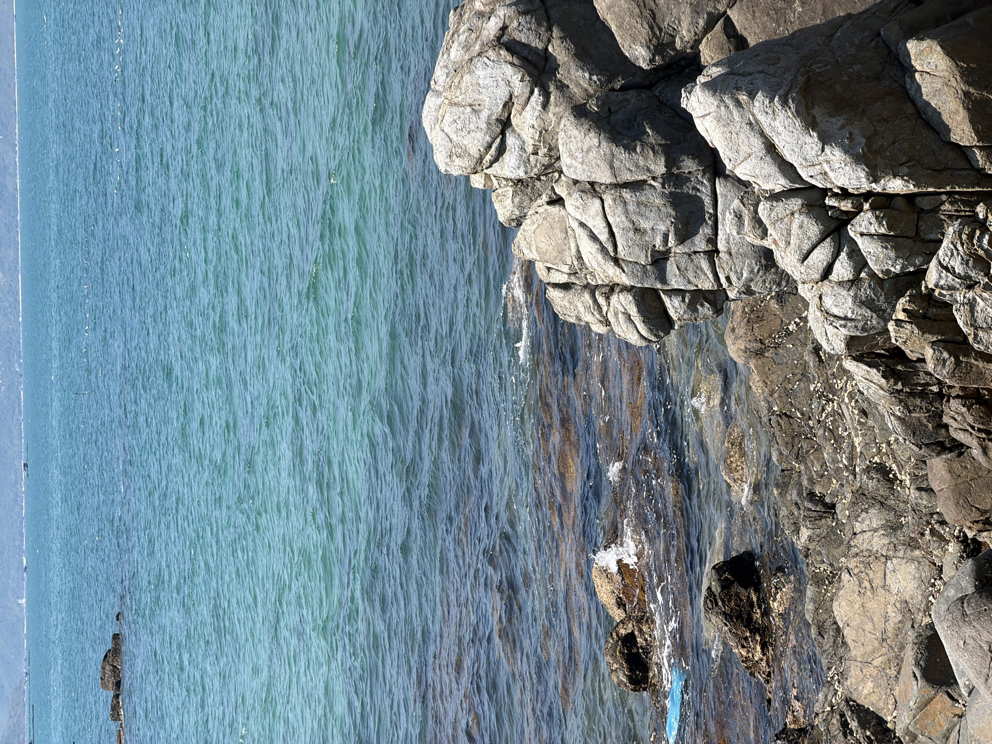 A scenic view shows the coast of Vĩnh Lương, Vietnam. Calm, turquoise water takes up most of the frame on the left side, extending to the horizon. Along the right edge of the image, a rocky coastline is present, with large, gray rocks forming the edge of the land. These rocks appear weathered and eroded by the sea, with some of the rock formations submerged in the water. There are no people visible in the scene.