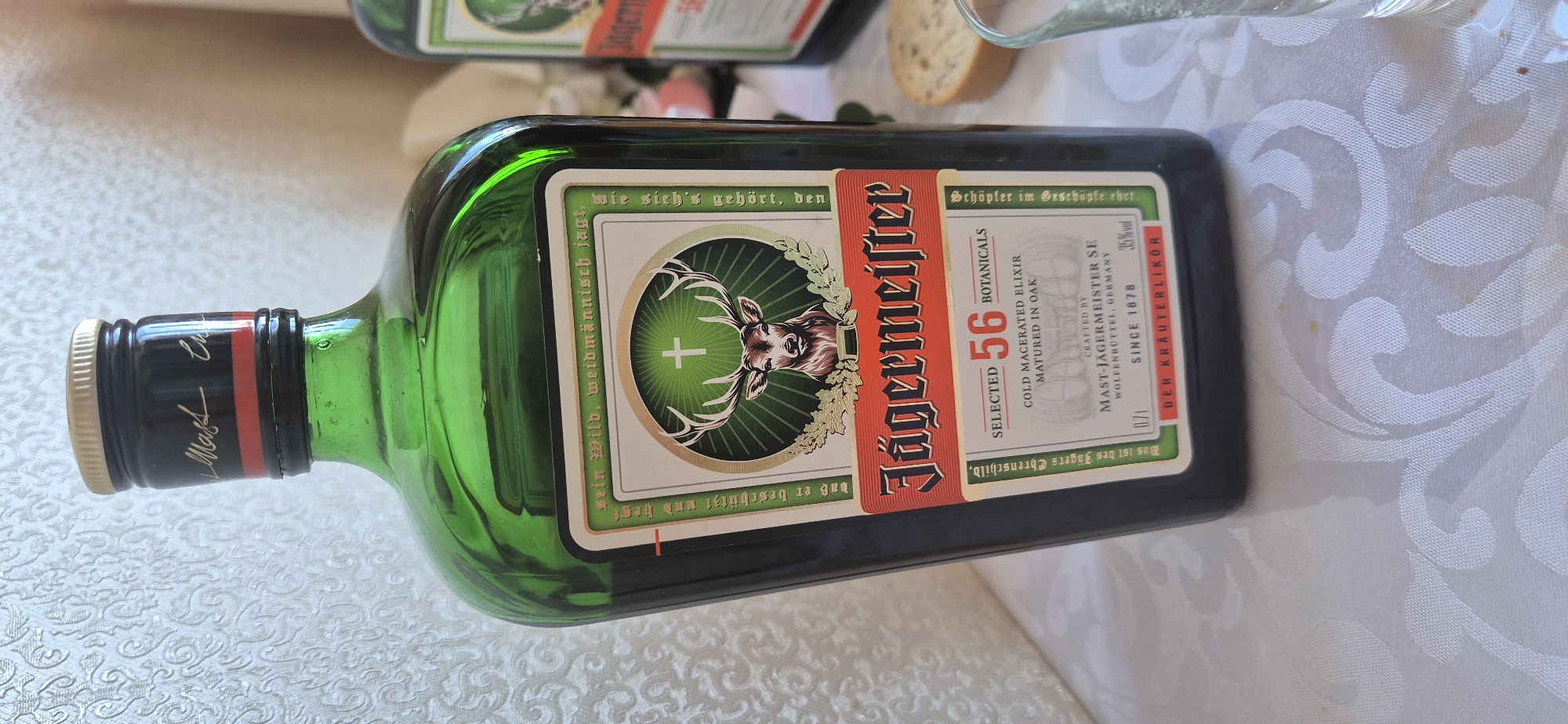A bottle of Jägermeister lies on its side on a patterned white tablecloth. The bottle is green with a black cap and a tan/gold label with the Jägermeister logo, which features a stag's head and the text "Jägermeister," "SELECTED 56 BOTANICALS," and "DER KRÄUTERLIKÖR." Another bottle is partially visible in the top-left corner. A piece of bread and a glass are also partially visible.