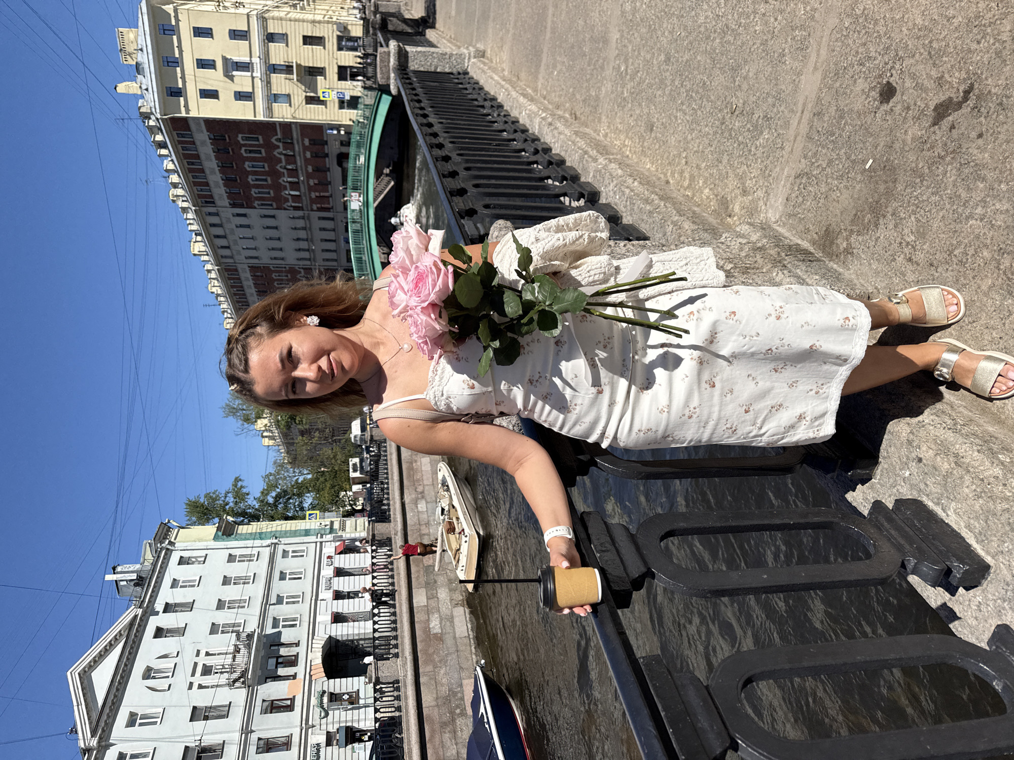 A female subject stands on a stone embankment next to a canal in an urban environment, likely in Saint Petersburg, Russia. She is positioned slightly to the right of the frame's center, facing directly towards the camera. She has light brown hair with braids, wears a white, floral-patterned midi-dress with thin straps, and light-colored open-toed sandals. In her left hand, she holds a disposable coffee cup with a dark straw, resting her arm on a black, ornate metal railing. On her left wrist is a white smart band or watch. Her right arm cradles a bouquet of approximately six to eight large, light pink roses with visible green stems and foliage. A cream-colored knitted garment is draped over her right arm, beneath the roses.
The background features a narrow canal with dark water, where two boats are visible: a dark blue covered boat on the left and a light-colored open motorboat with a single person in a red shirt on the right, further down the canal. The canal is bordered by a stone embankment and the black metal railing where the subject leans. Across the canal, a row of multi-story, historic European-style buildings lines the waterfront. These buildings exhibit varying facades, including a light blue-grey structure with white trim and a prominent pediment on the left, and a longer building with red brick and yellow/cream sections on the right. A green arched bridge spans the canal in the mid-distance. The sky is clear and bright blue, with several utility wires visible overhead. The ground in the foreground is a textured light grey pavement.