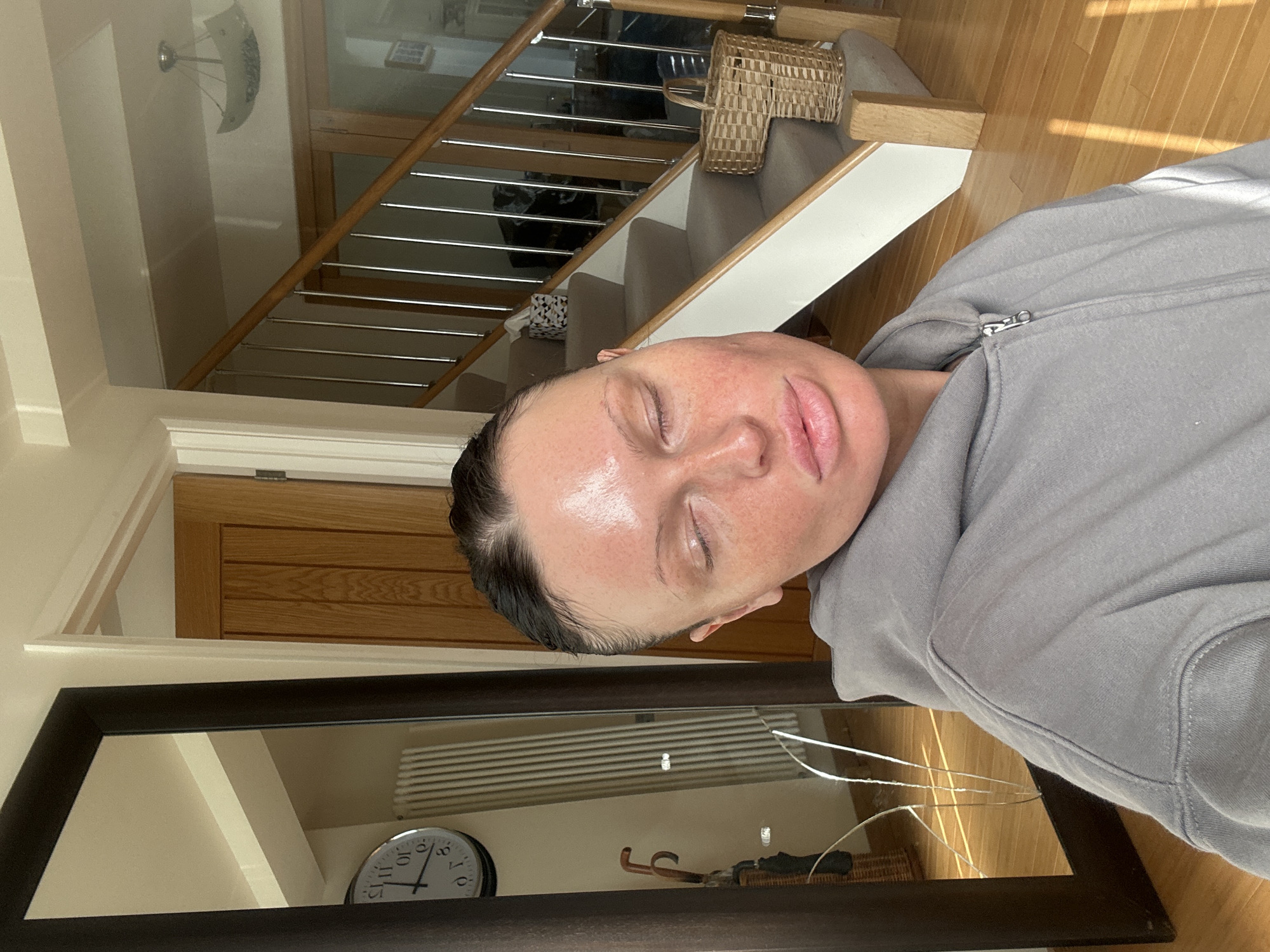 An individual is depicted in an interior residential setting, consistent with a dwelling in the United Kingdom, potentially within the Wealden district. The subject, an adult with dark hair pulled back, has closed eyes and a reddish, shiny appearance on the face, suggesting a facial treatment. They are wearing a grey zippered hooded sweatshirt. To the left, a large, dark-framed mirror reflects a white panel radiator, a round wall clock with Roman numerals, and a wicker umbrella stand holding an umbrella. To the right, a carpeted staircase with wooden banisters and metal spindles ascends. A woven basket rests on a lower stair, and a patterned tissue box is visible on an upper stair. Wooden doors are present in the background, along with a ceiling light fixture. Streaks of sunlight illuminate the wooden floor in the foreground.
