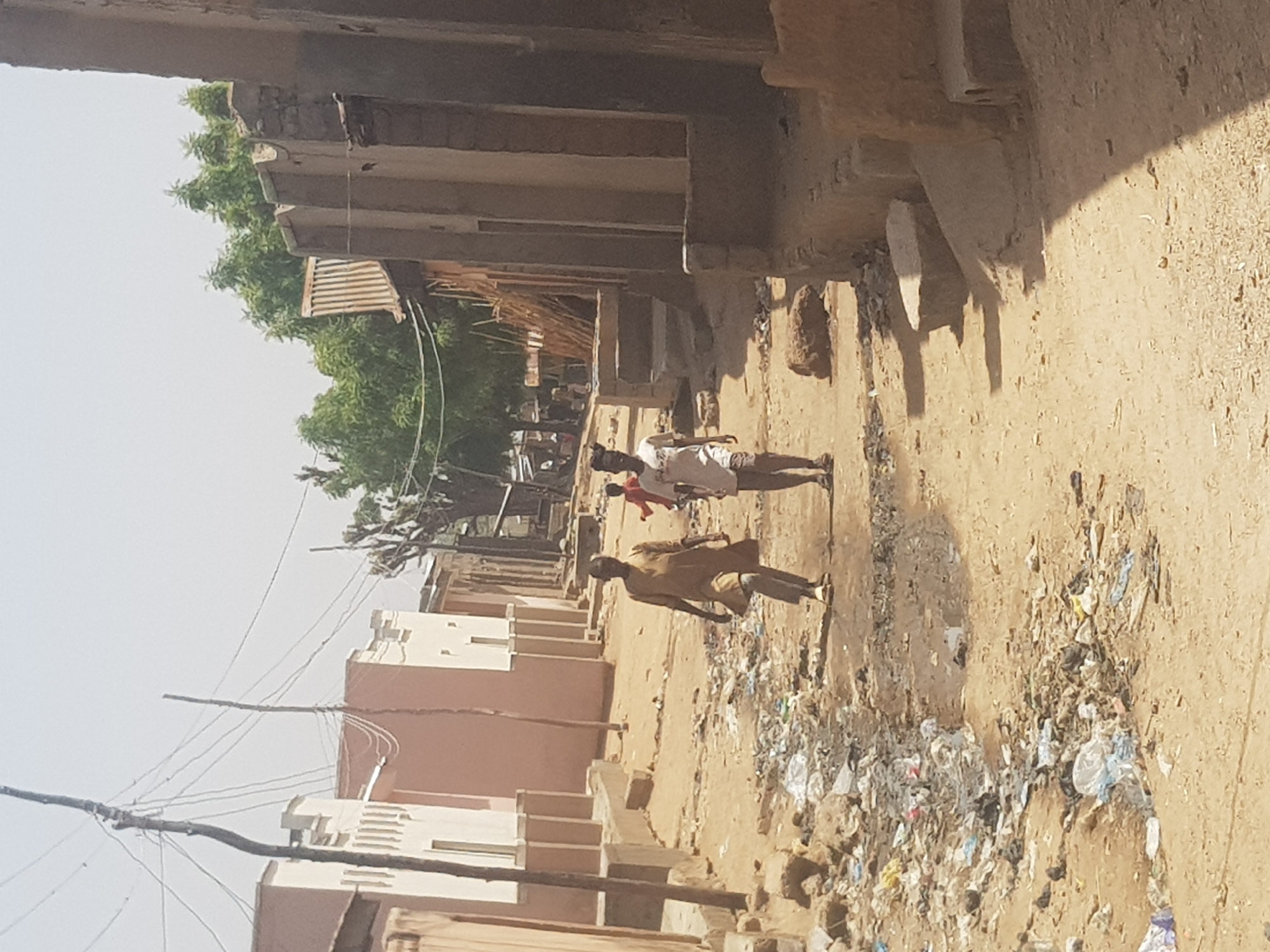 The image depicts a street scene in Dutsinma B, Nigeria. The street is unpaved and covered in litter and debris. There are two people walking down the street. One person is wearing a brown shirt and the other is wearing a white shirt with a red top. A child is also visible walking with the person wearing the white shirt. The buildings on either side of the street are mostly made of mud and brick. Overhead power lines are present.