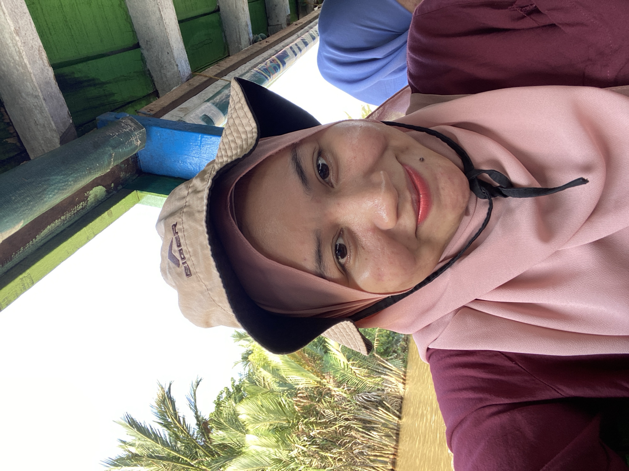 A medium shot selfie features a woman wearing a pink hijab and a tan bucket hat with a black brim. She is wearing a maroon shirt. Her right eye is visible and she is smiling. Behind her, palm trees and water are visible, indicating an outdoor environment, likely a river or canal. The background suggests a tropical location. To the right, a portion of another person wearing a blue hijab is visible. The scene appears to be on a boat, with wooden structures overhead. The overall impression is a personal snapshot taken during an excursion.