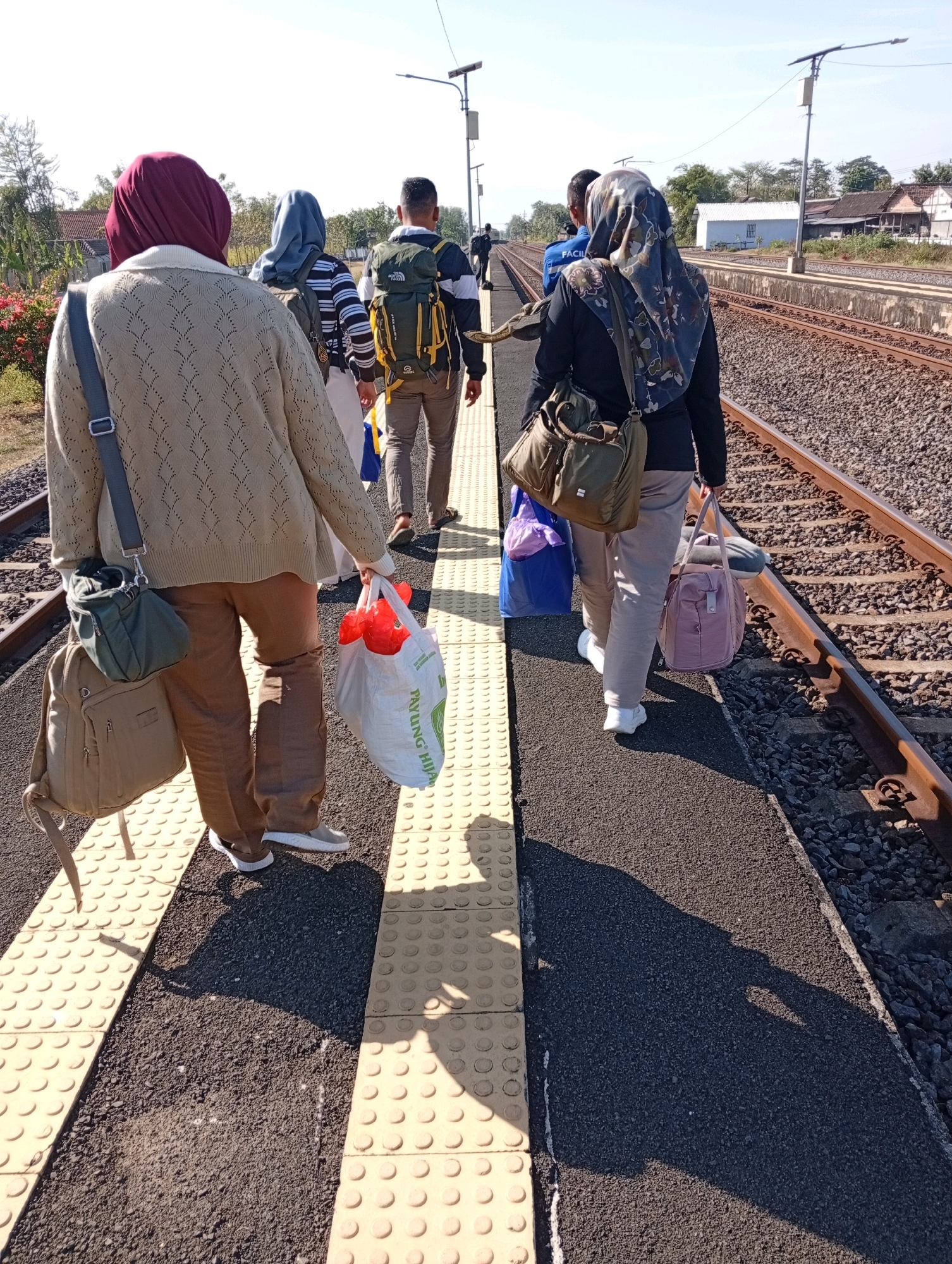 In Tebon, Indonesia, a group of people walk along a train platform. The platform has a yellow tactile paving on the edge. The train tracks are to the right of the platform, covered with ballast. Sunlight casts long shadows on the ground. On the left, a person wearing a maroon hijab and a tan patterned cardigan walks away from the camera, carrying a tan backpack and a green bag. They are also holding a white plastic bag containing red items. Behind them, another person in a blue hijab, wearing a striped shirt and carrying a backpack, walks forward. To their right, a man in a backpack and a patterned shirt walks away. Further ahead, a person in a dark hijab and black top carries a patterned bag and a pink duffel bag. The background shows trees, a white building, and houses. The sky is clear and blue.