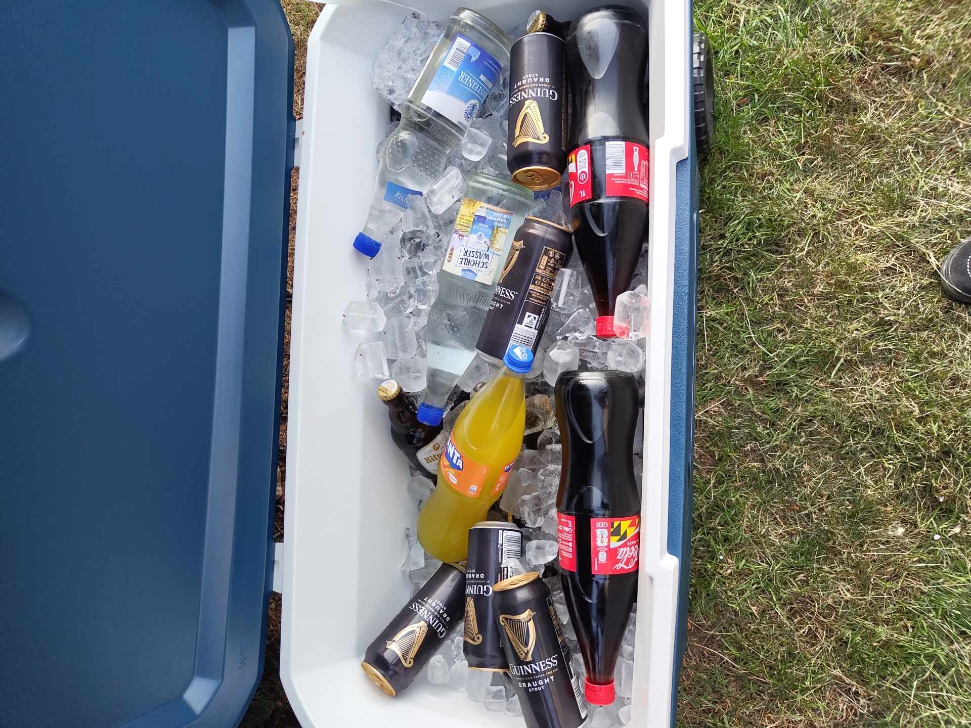 The image shows a blue and white cooler filled with ice and various beverages. The cooler appears to be outdoors on a grassy surface, based on the visible background. Inside the cooler, there are bottles of water (including one labeled "STEINER"), glass bottles of "SCHORLE WASSER," a yellow Fanta bottle, bottles of Bitburger beer, and Coca-Cola bottles. Additionally, there are several cans of Guinness Draught stout. The cans and bottles are surrounded by ice. A portion of a shoe is visible on the right side of the image. The image contains the words "Schmelz, Germany" on one of the labels of the bottled water.