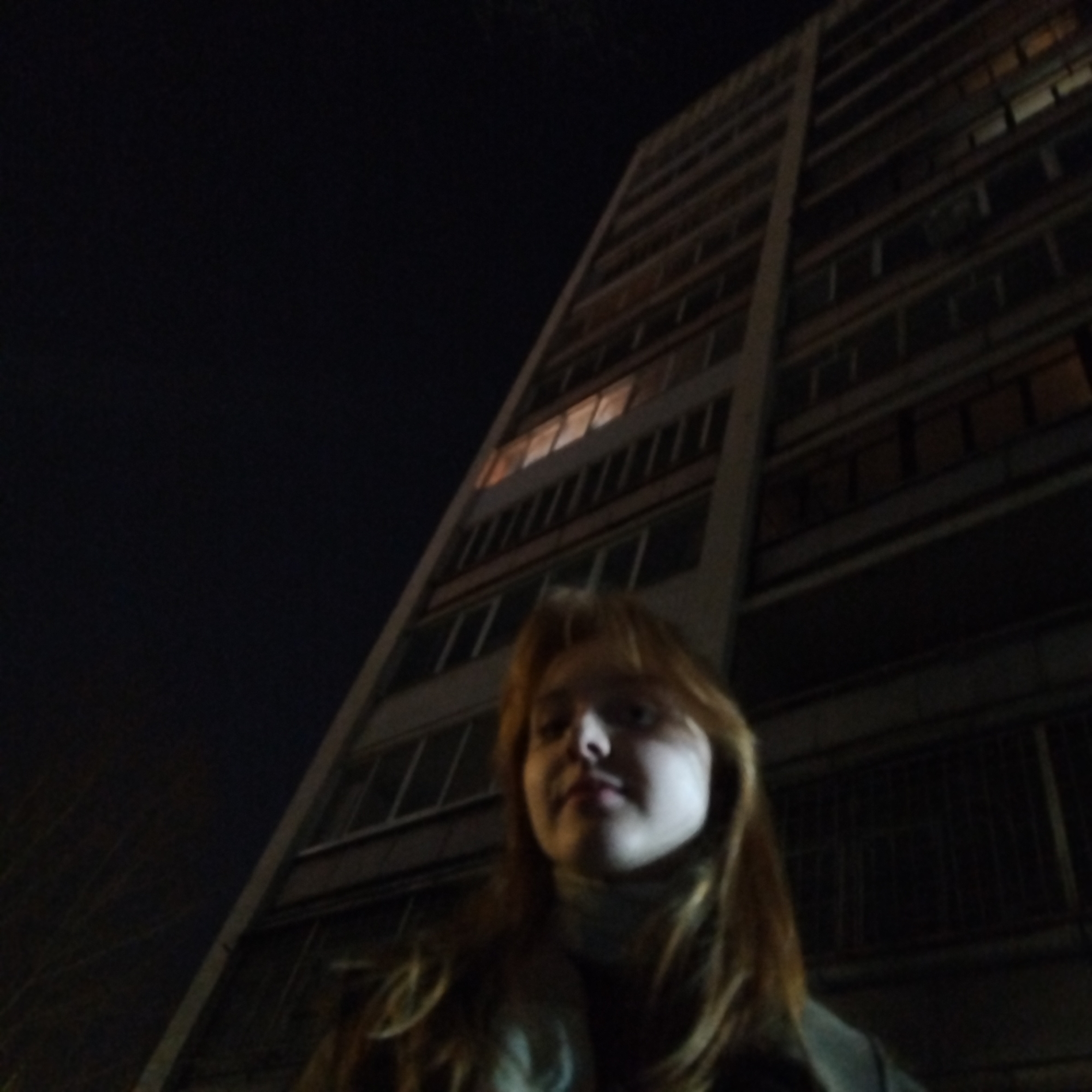 A woman with long brown hair, wearing a collared shirt, stands in the foreground, looking slightly upward into the camera. Behind her is a tall, multi-story building with a rectangular shape. The building has many windows arranged in a grid-like pattern, with some lit from within. The sky is dark, suggesting it is nighttime. Trees are visible on the left-hand side. Based on the architectural style, the building is likely located in Yekaterinburg, Russia.