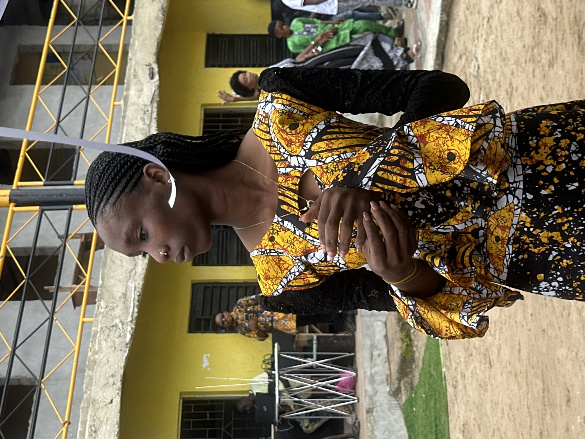 In Badagry, Nigeria, a young woman with braided hair and a nose ring stands outside a yellow, two-story building with a concrete foundation. She wears a vibrant, patterned dress with a cutout at the neckline, black lace sleeves, and a flared skirt. A white ribbon is attached to her hair. Her hands are clasped in front of her. In the background, other individuals are visible. To the left, a person is seated at a table with a laptop. Further back, a man in a matching patterned shirt stands near a doorway, and other people are milling about. The ground is sandy, and a grassy area is present to the left. The building features yellow walls, dark-framed windows, and a yellow metal railing on an upper level.