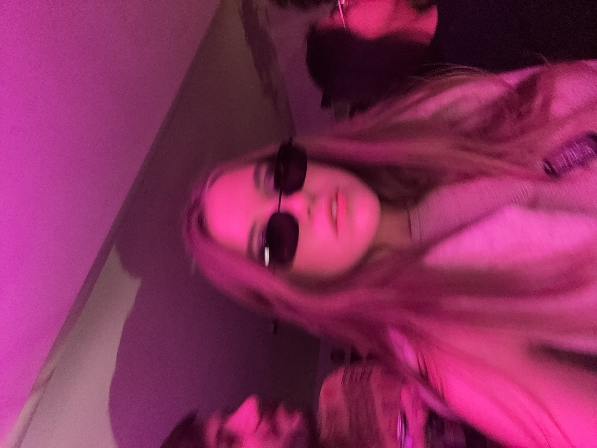 A blurry image presents two individuals, one female wearing dark sunglasses and a light-colored, possibly pink, sweater, and another individual with dark hair. The setting appears to be indoors under pink lighting. No specific details indicate a location in Mexico City.