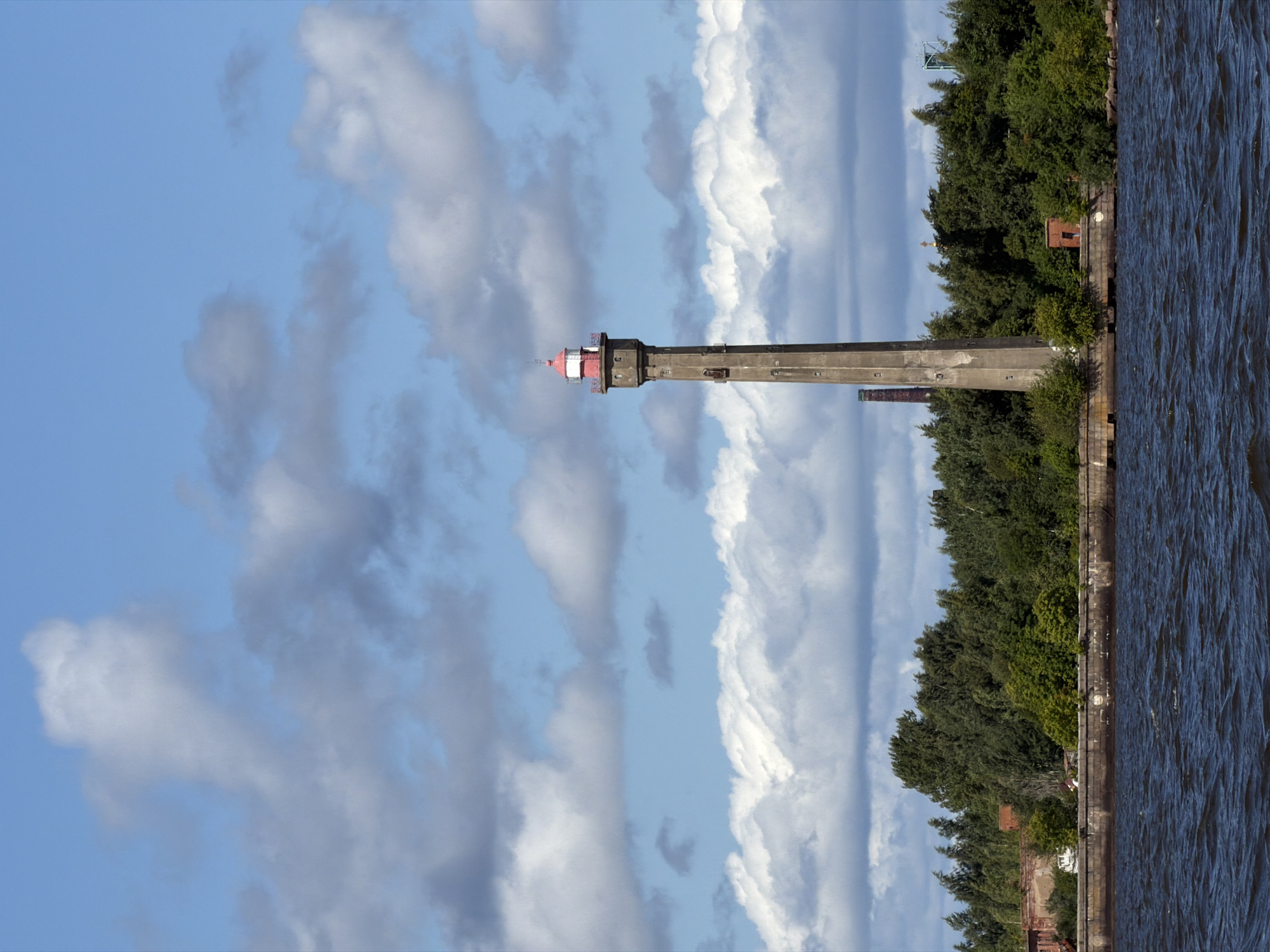 A tall, concrete lighthouse with a red cap stands prominently on the shore of the Gulf of Finland in Lomonosov, Russia. The lighthouse, constructed of large, square blocks, features numerous small windows evenly spaced along its height, and an attached metal ladder on its left side. The top of the lighthouse is capped with a red lantern room. Behind the lighthouse, a dense line of green trees runs along the waterfront, with some older brick buildings and a brick chimney visible amongst them. The water in the foreground is a dark, choppy blue, reflecting the bright sky above, which is dotted with dramatic white and gray clouds.