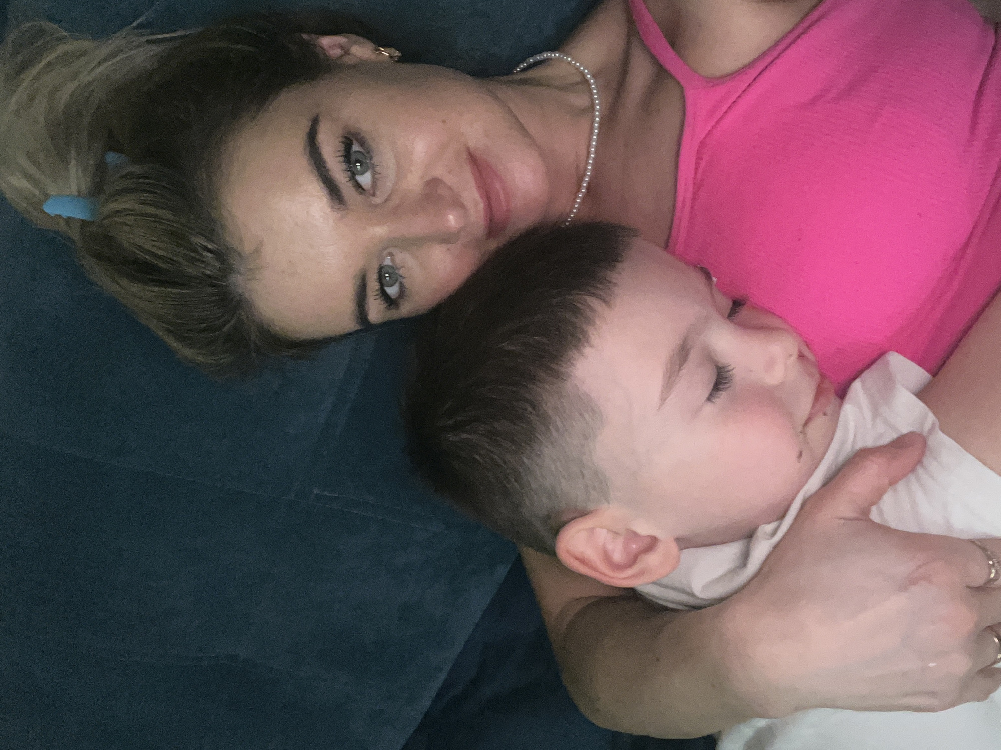A woman wearing a pink tank top and a pearl necklace is lying on a dark blue upholstered surface, possibly a couch or bed. She is looking at the camera with a slight smile. A young child is nestled in her arms, appearing to be asleep, and is wearing a white shirt. The woman's arm is around the child. A blue hair clip is visible in the woman's hair. The scene appears to be indoors. The location, Chaikovsky, Russia, cannot be confirmed from the visual information.