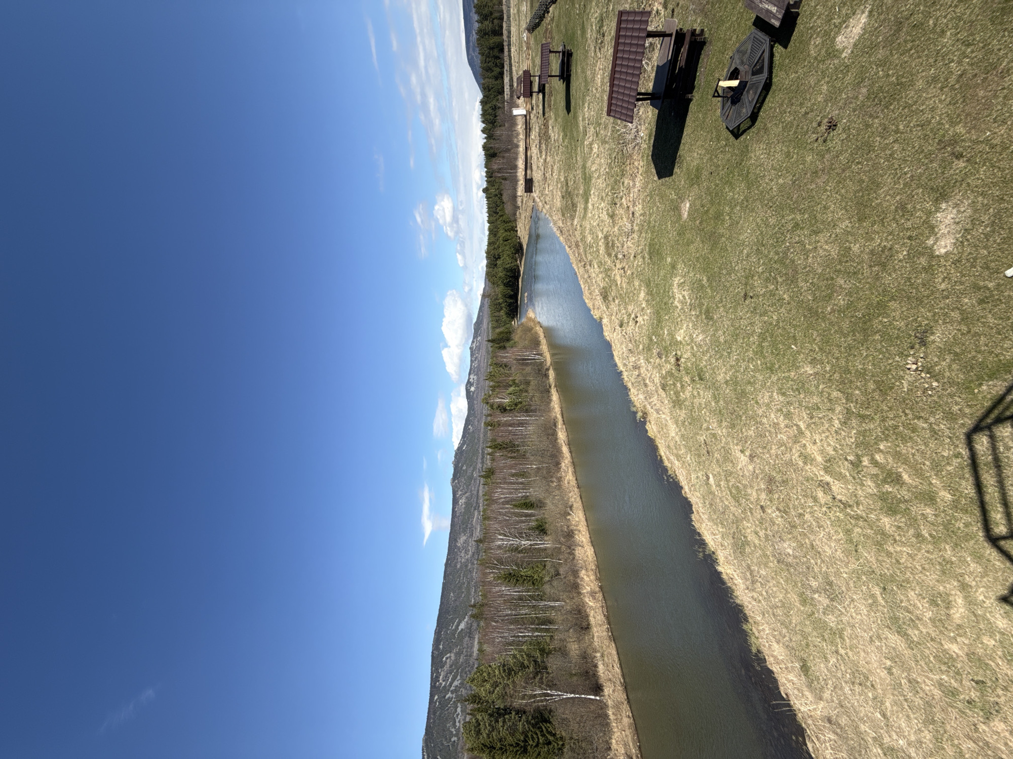 The image displays a panoramic outdoor scene, likely a recreational area in Kordonnyi, Russia. A calm, narrow body of water, possibly a canal or river, horizontally bisects the middle ground of the landscape. On the closer bank, covered in a mix of dry and emerging green grass, several wooden picnic tables with benches are visible, some featuring dark, shed-like roofs. An octagonal fire pit or grill structure is also present on this bank.
The opposite bank is lined with a dense growth of trees, appearing to be a mix of coniferous and deciduous species, with many deciduous trees still bare, suggesting early spring. Beyond this treeline, a range of mountains rises gently in the distance, partially obscured by the trees. The upper portion of the image is dominated by a vast, clear blue sky dotted with a few scattered white clouds.
The scene suggests a tranquil outdoor setting designed for leisure activities. The picnic tables and fire pit indicate the area is suitable for gatherings, picnics, or barbecues, though no people or active events are depicted. The time of day appears to be sunny midday or early afternoon, with bright illumination and distinct shadows cast by the tables. The weather is clear, enhancing the serene atmosphere of this natural and developed recreational space. No visible text is present in the image.