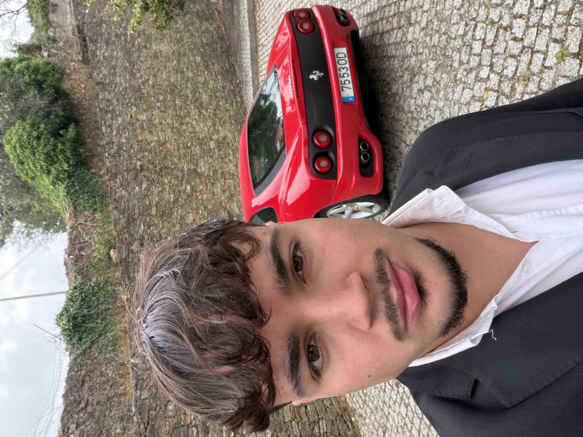 A young man wearing a black suit jacket and a white collared shirt takes a selfie next to a red Ferrari. The license plate of the car reads "7553OD" and has a Portuguese flag on the left side. The setting appears to be outdoors, with a stone wall covered in green vegetation behind the car. The ground is paved with cobblestones. Based on the license plate and architecture, the location appears to be in Vila Real, Portugal.