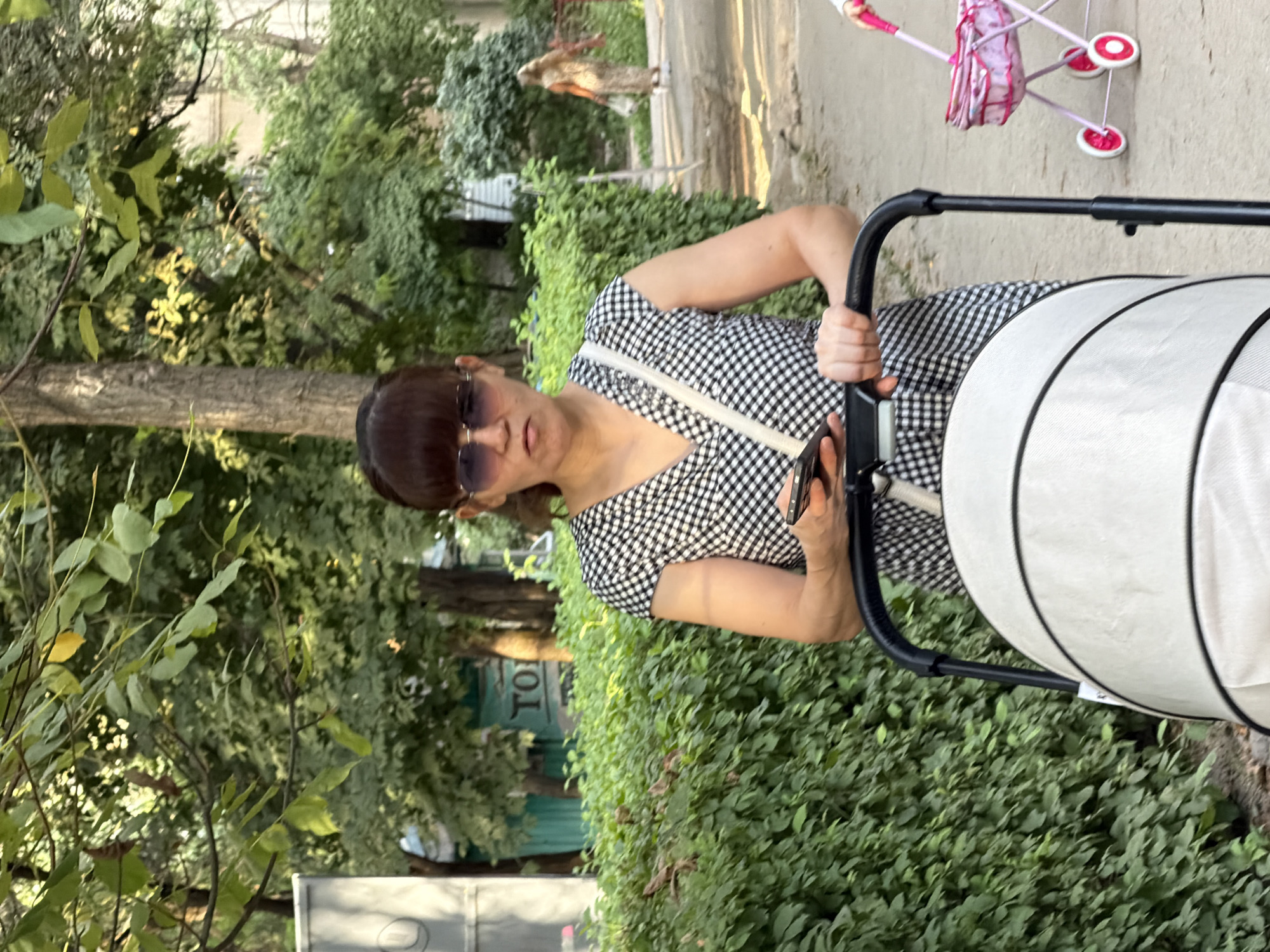 In an outdoor urban setting, a woman with dark hair pulled back and bangs, wearing sunglasses and a black and white checkered dress, stands holding a smartphone in her left hand and gripping the handle of a white baby stroller with her right. She is looking directly at the camera with a neutral expression. Behind her, a dense green hedge forms a backdrop, and further beyond, trees and foliage suggest a park or residential area. To the right of the woman, a small pink toy stroller with pink wheels is visible. In the background, a woman in a long, patterned dress walks away from the camera. The ground appears to be a dirt or gravel path.