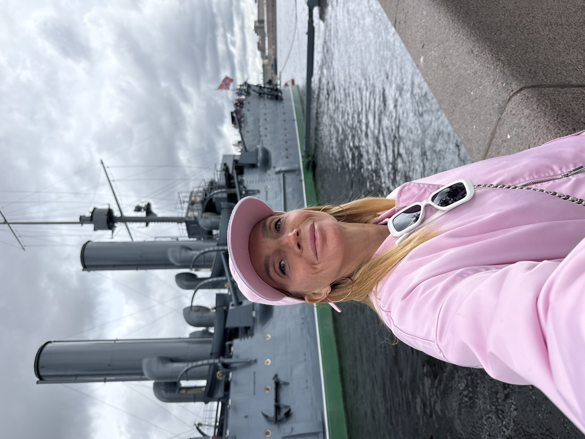 A woman is taking a selfie in front of the Cruiser Aurora in Saint Petersburg, Russia. The woman wears a pink jacket and cap, with white sunglasses atop her head. The Cruiser Aurora, a Russian protected cruiser, is docked nearby. The water is calm. The sky is overcast.