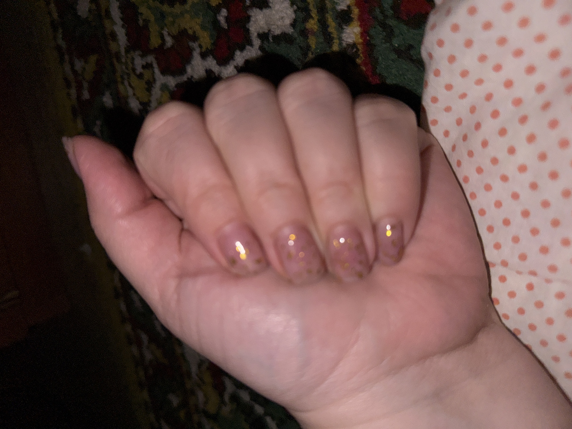 A close-up, overhead shot of a person's left hand, clenched into a fist, rests on a pink fabric with white polka dots. The hand is positioned in the lower right quadrant of the frame. The nails are painted with a sheer, light pink polish adorned with small gold flecks. The background is dark and out of focus, featuring what appears to be a patterned rug with muted colors. The lighting suggests it is indoors, likely in a residential setting. The specific location in Ketovo, Russia, is not discernible from the visual information provided in the image.