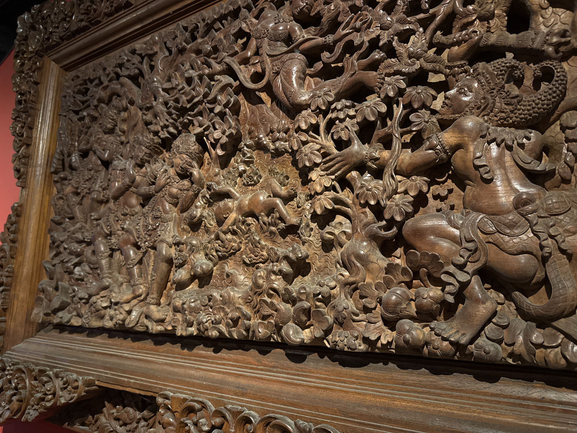 This is a close-up, angled shot of a highly detailed wooden relief carving, likely a decorative panel or part of a larger architectural element. The carving depicts a dense, dynamic scene filled with human figures, fantastical creatures, and intricate floral motifs.
The subjects are predominantly human-like figures in various poses. Many appear to be engaged in some form of narrative action. Some figures are standing, others are seated or in mid-motion, their bodies contorted to fit within the carved space. There are also elements that suggest mythical beings or deities, such as the scaled tail of a creature on the right side of the frame, and the ornate headdresses and jewelry worn by some figures. The scene is densely packed with foliage, including detailed carvings of flowers and leaves, creating a lush, organic environment.
The overall impression is one of activity and storytelling, though the specific narrative is not immediately clear without further context. The figures' expressions are difficult to discern due to the carving's depth and texture, but some appear to be gesturing or interacting with each other. The time of day is suggested by the warm lighting and shadows that highlight the depth and texture of the wood, casting dramatic contrasts across the relief. The setting appears to be indoors, likely a museum or gallery, given the lighting and the way the carving is presented on a reddish wall.
Notable details include the fine craftsmanship evident in the rendering of muscles, hair, clothing folds, and intricate patterns. The texture of the wood itself, with its grain and age, adds to the visual richness. The base of the carving features a more geometric, patterned border, contrasting with the organic scene above. There is no visible text in the image. The style of the carving suggests an Indonesian origin, possibly Bali, given the location context provided.