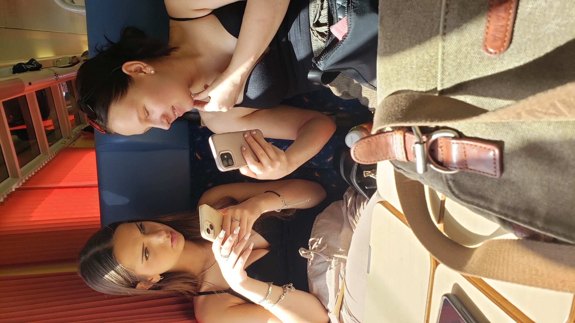 Two young women are seated next to each other, possibly on a train, looking at their smartphones. The woman on the left, with long brown hair, wears a black tank top and light-colored pants. She holds a gold-colored smartphone in her left hand and is looking at it with a serious expression. She has several bracelets on her left wrist and rings on her fingers. The woman on the right, with dark hair pulled back, wears a black tank top and is looking down at her phone, which is also gold-colored. She has earrings in her ears and her right hand is near her chin. Between them, a brown bag with leather accents is visible in the foreground, partially obscuring a table. Behind them, the orange curtains of what appears to be a train carriage are illuminated by sunlight. The environment suggests they are indoors, possibly traveling. The context provided about Ladce, Slovakia, is not visually represented in the image itself.