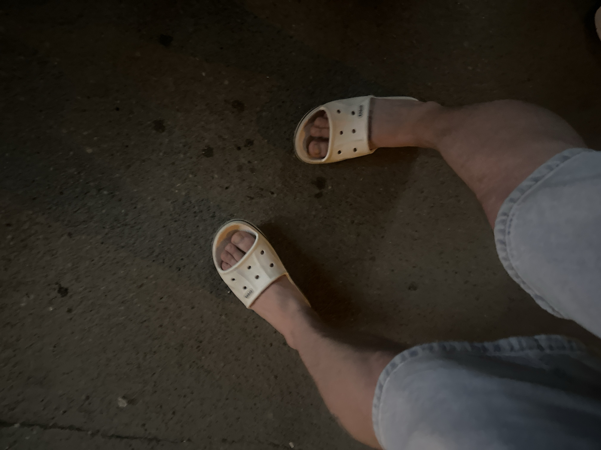 A top-down view captures a person's legs and feet wearing light-colored slide sandals. The individual is standing on a dark, textured surface that resembles asphalt or concrete. The person is wearing light-colored denim shorts. The location is not specified in the picture.