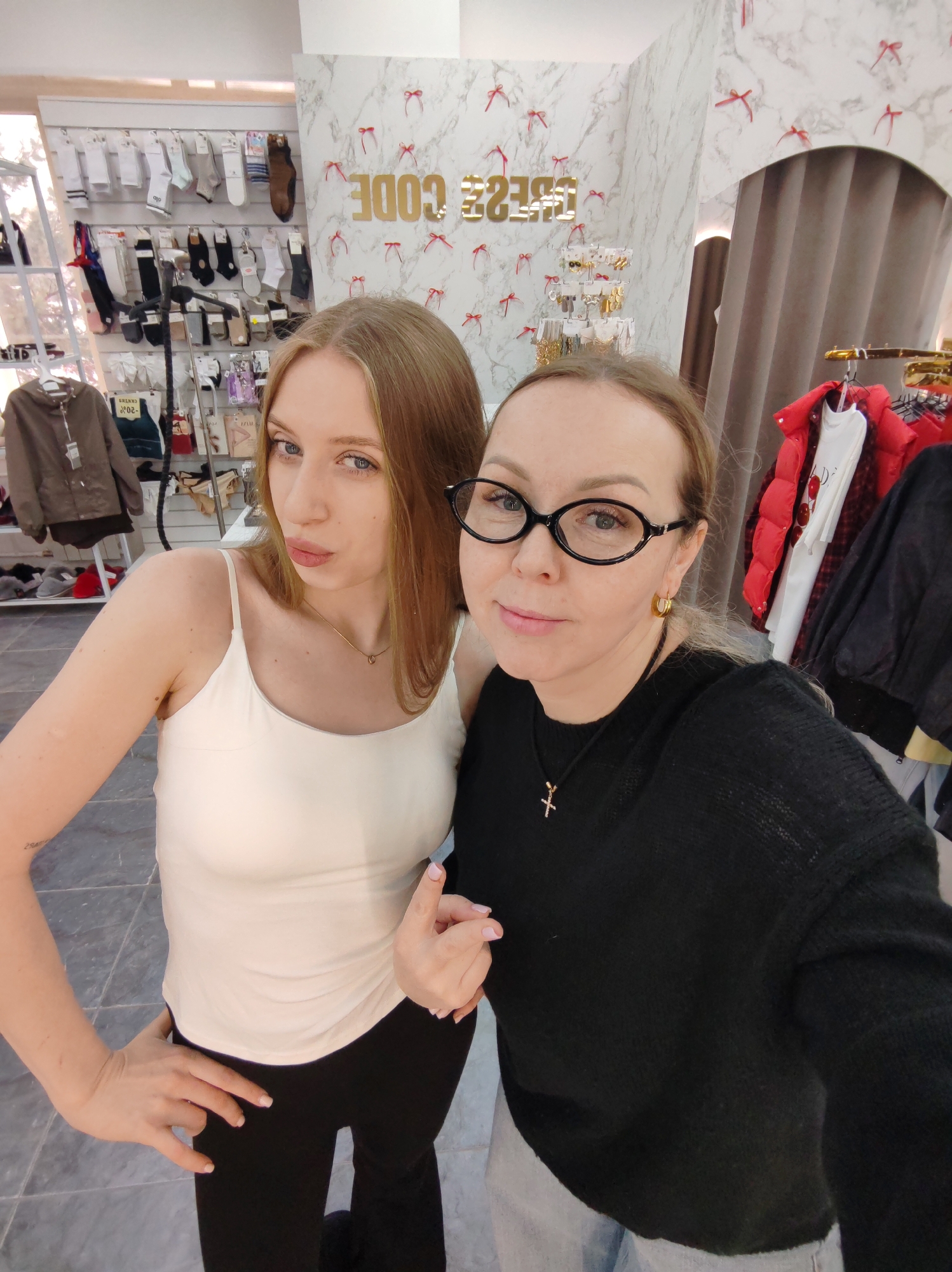 Two adult women are captured in a selfie within a retail clothing store named "DRESS CODE" in Chaikovsky, Russia. The woman on the left has long blonde hair, is wearing a light-colored spaghetti strap top, dark pants, and a delicate gold-colored necklace. She poses with her right hand on her hip and her lips slightly pursed. A faint tattoo is visible on her left forearm. The woman on the right, taking the photograph, has blonde hair, wears black oval-shaped glasses, gold hoop earrings, a black long-sleeved sweater, and a gold cross pendant necklace. Her left hand is partially visible with the thumb extended. The store environment features white marble-patterned wallpaper adorned with small red bow ties and the gold-lettered "DRESS CODE" sign, which appears reversed due to the selfie angle. Merchandise visible includes shelves stocked with socks and hosiery, clothing racks displaying various garments such as a red puffer jacket over a white t-shirt with cherries, and a stand with jewelry. A brown dressing room curtain is partially visible on the right. The floor is tiled with gray squares.