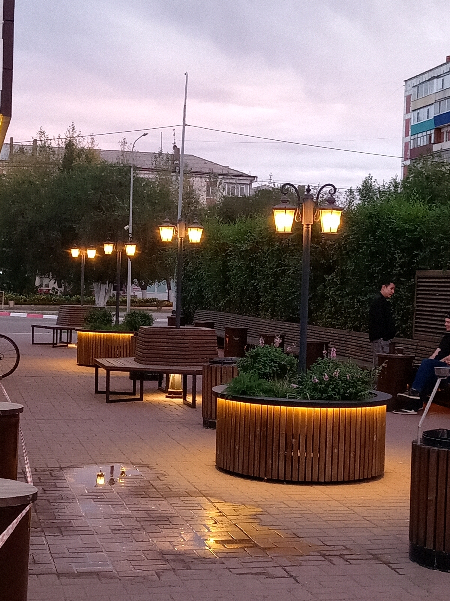 The image depicts an outdoor scene in the city of Rudny, Kazakhstan. The time appears to be dusk, with a cloudy sky casting a soft, ambient light. The foreground is a brick-paved walkway with a puddle reflecting some of the surrounding lights.
Several modern, wooden planters with integrated yellow lighting are visible, adding a warm glow to the area. Each planter is topped with greenery. Adjacent to the planters are wooden benches, some with dark metal frames, designed for seating. A bicycle wheel is partially visible on the left side of the frame.
Streetlights with a classic design, featuring multiple lamps emitting a warm yellow light, are positioned along the walkway. In the background, there are buildings, including a multi-story apartment building with colorful balconies and some lower-rise structures. Dense green bushes and trees form a backdrop behind the seating areas.
Two individuals are present in the scene. One person, a male, is standing near a planter and looking away from the camera, appearing to be in conversation. Another person, also appearing to be male, is seated on a bench in the mid-ground, also facing away from the camera. The overall impression is a public space designed for relaxation and outdoor gathering, illuminated by artificial lighting as daylight fades.