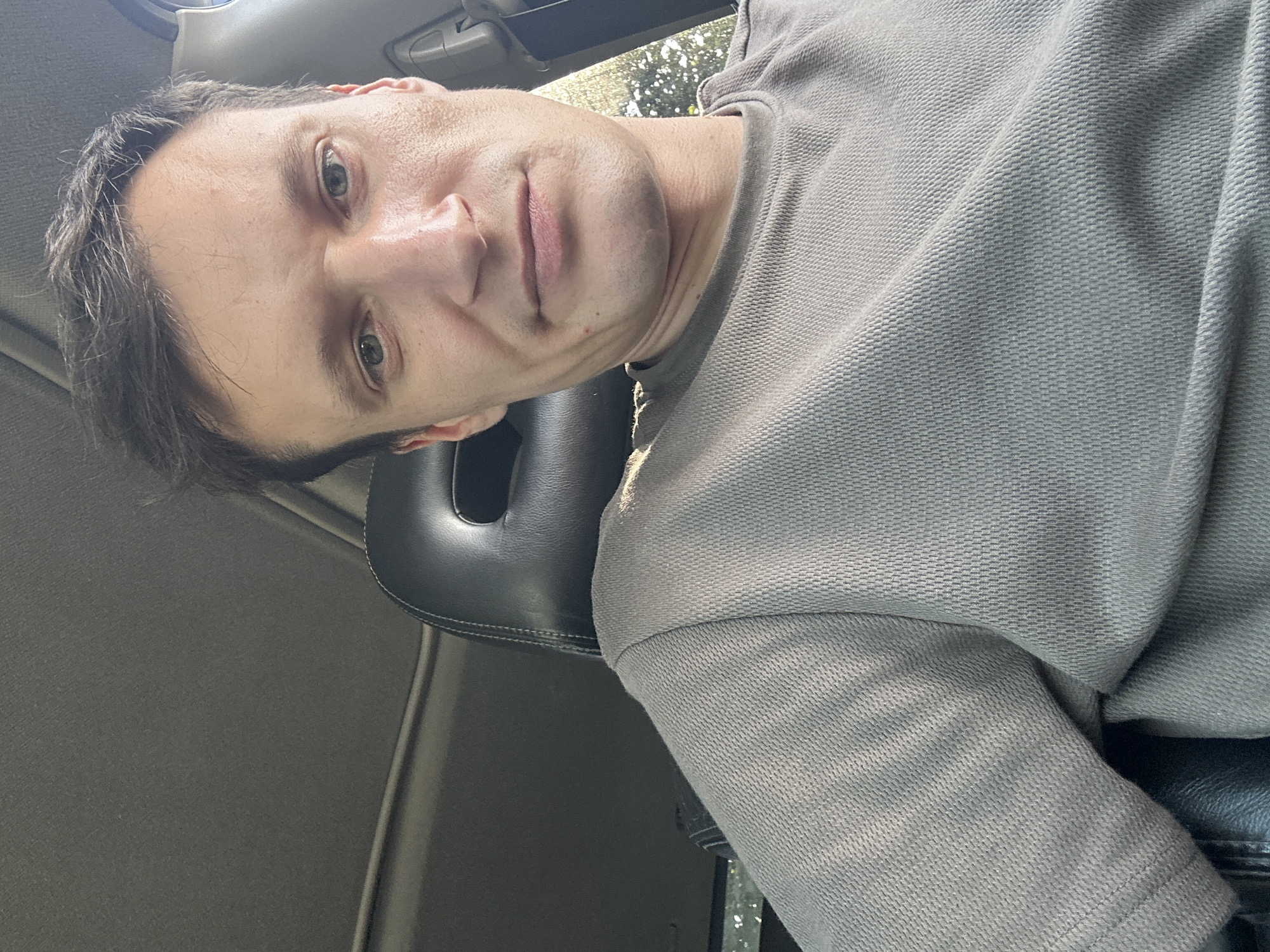 A medium-shot, selfie-style photo shows a white male with short brown hair, light blue eyes, and fair skin. He is wearing a grey t-shirt and is seated inside a vehicle. The background includes the interior of the car, with a black headrest and a sunroof visible. Sunlight filters into the car, illuminating the subject and the surrounding environment, with foliage visible through a side window. There are no discernible landmarks indicating the specific location of Shatava, Ukraine.
