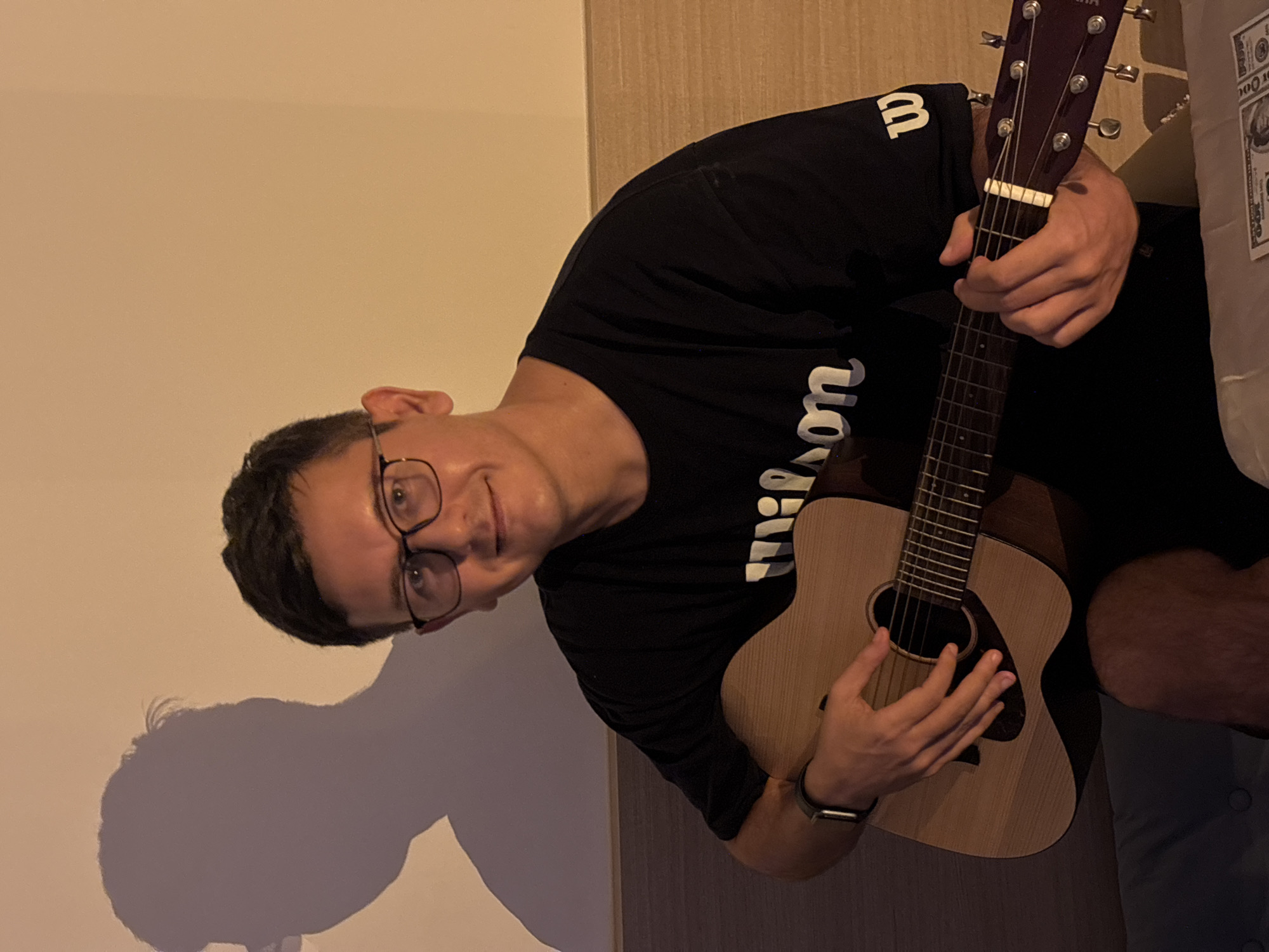 A man with dark hair and glasses is sitting down and playing a guitar. He is wearing a black t-shirt with white writing that says "Wilson." There is a shadow of the man on the wall behind him. The room appears to be dimly lit with warm lighting. The background wall is a light tan color. There are what appear to be some US dollar bills on a table in the foreground.