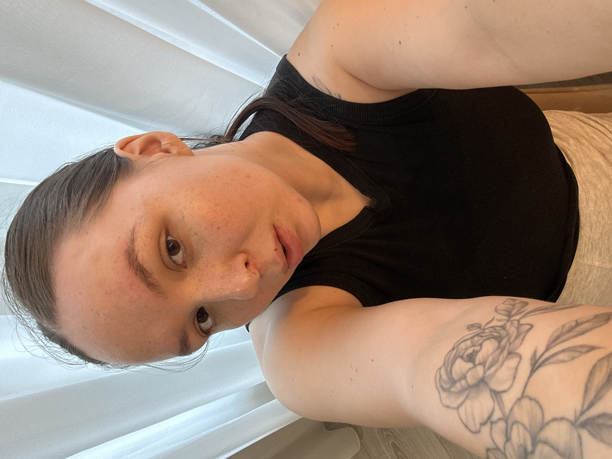 This is a close-up, selfie-style photograph taken indoors. The subject is a person with fair skin, freckles, and dark hair pulled back. They are wearing a black, ribbed tank top and light-colored bottoms. A tattoo of a flower, possibly a peony, is visible on their left arm. The background consists of white curtains or blinds and a wooden floor. The lighting suggests it is daytime. The person's head is tilted, and their gaze is directed towards the camera, with a neutral or slightly relaxed expression. There is no visible text in the image. The context provided places the location in Chaikovsky, Russia.