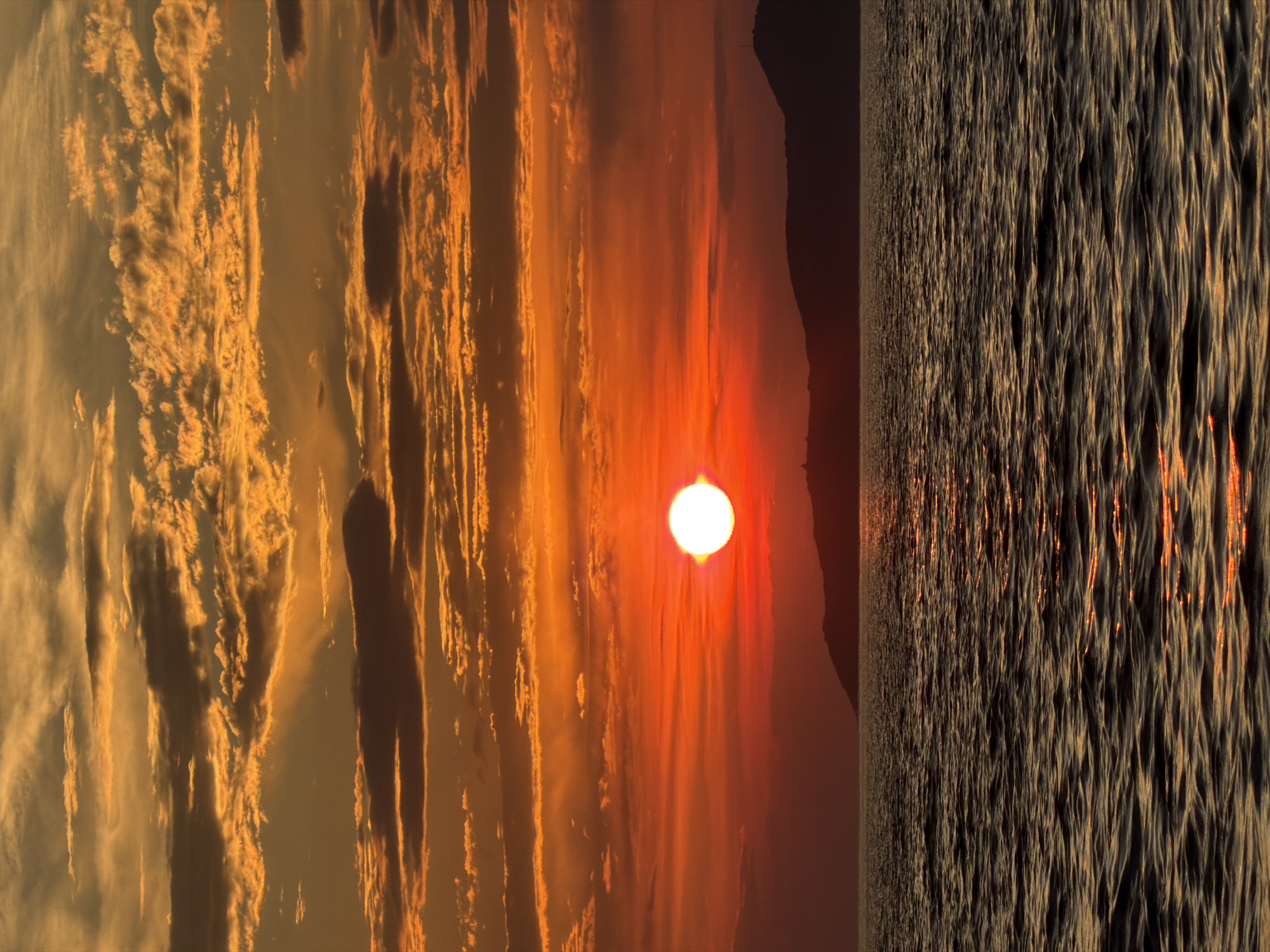 The image captures a dramatic sunset over the sea, with the sun appearing as a bright, fiery orb. The sky is filled with vibrant clouds, painted in hues of orange, gold, and red, creating a striking contrast against the darkening landscape. To the right, the choppy surface of the water reflects the warm tones of the sunset, with streaks of light shimmering across its textured surface. A dark silhouette of a hill or landmass is visible in the background, adding depth to the scene. The overall impression is one of serene beauty and the awe-inspiring power of nature. The orientation of the image is vertical. No people or text are visible in the image. The scene likely depicts the sunset at Petrovac centar, Montenegro.