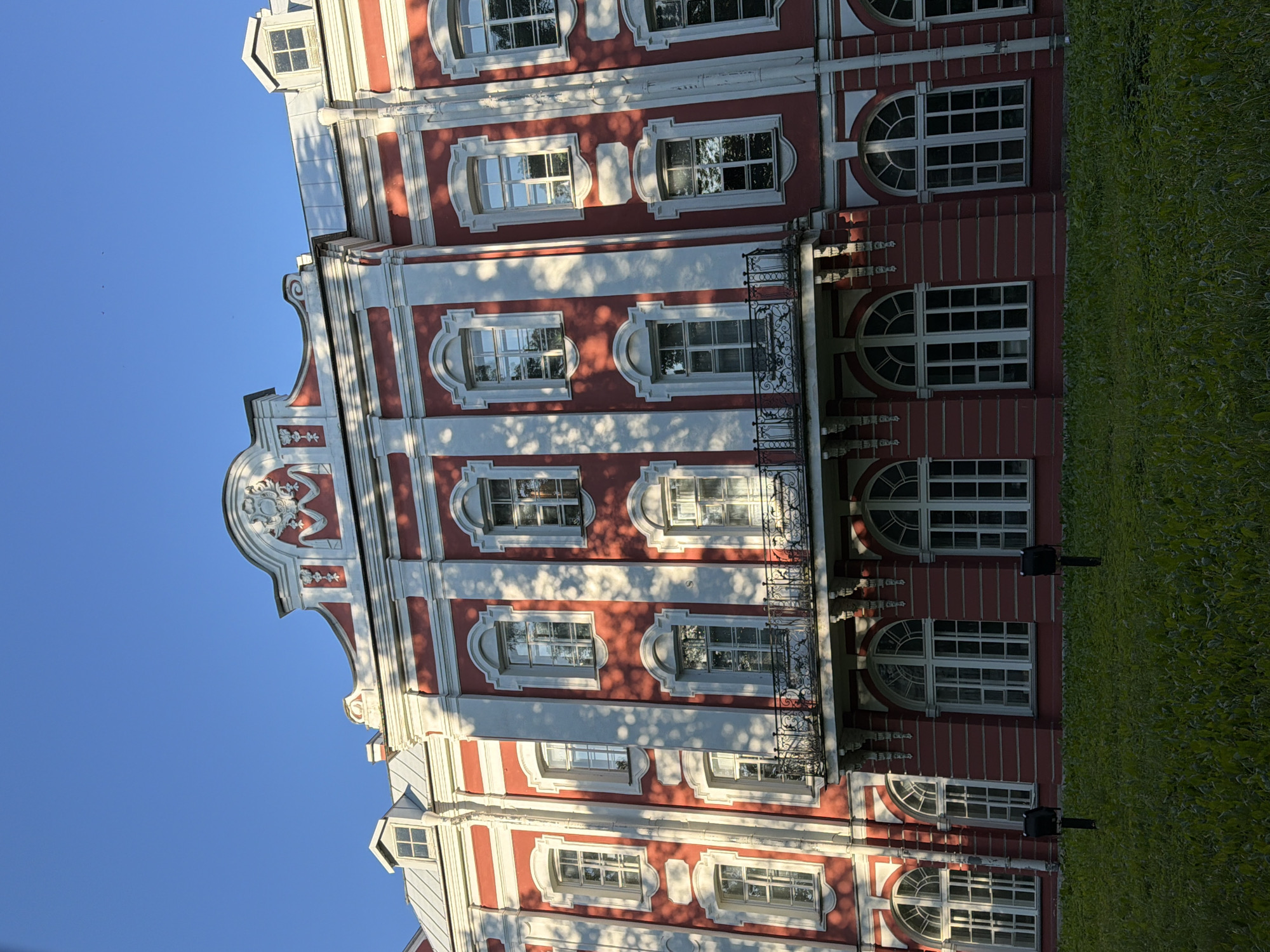 This is a photograph of the Catherine Palace (also known as Tsarskoye Selo) in Pushkin, a town within the federal city of Saint Petersburg, Russia. The image captures a section of the palace's red facade, adorned with white architectural details. Numerous windows, some arched and others rectangular, punctuate the building. A decorative balcony with an ornate railing extends from the middle floor. The upper part of the building features a pediment with intricate ornamentation. Sunlight casts dappled shadows from trees onto the palace walls. In the foreground, a neatly manicured lawn stretches across the scene, with two dark, cylindrical bollard lights positioned on the grass. The sky above is a clear, deep blue.