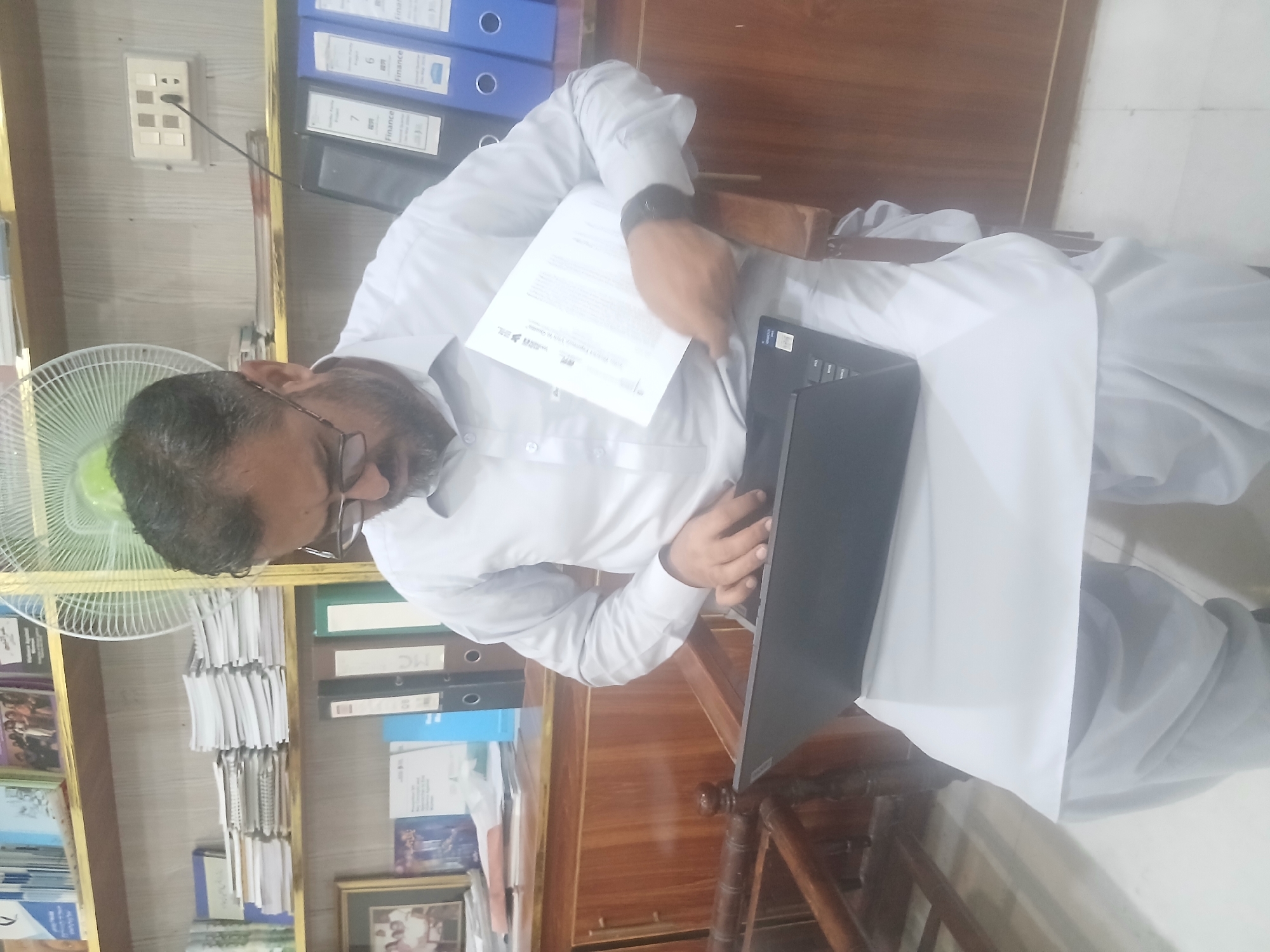 A man wearing glasses, a long-sleeved white shirt, and traditional shalwar kameez is seated in a room. He is holding a piece of paper in one hand and using a laptop on a wooden desk or table in front of him. Behind him is a shelf containing files, books, and a white electric fan. The image seems to be taken in an office. The location mentioned, Quetta City Tehsil, Pakistan, can be considered the possible context for the setting.