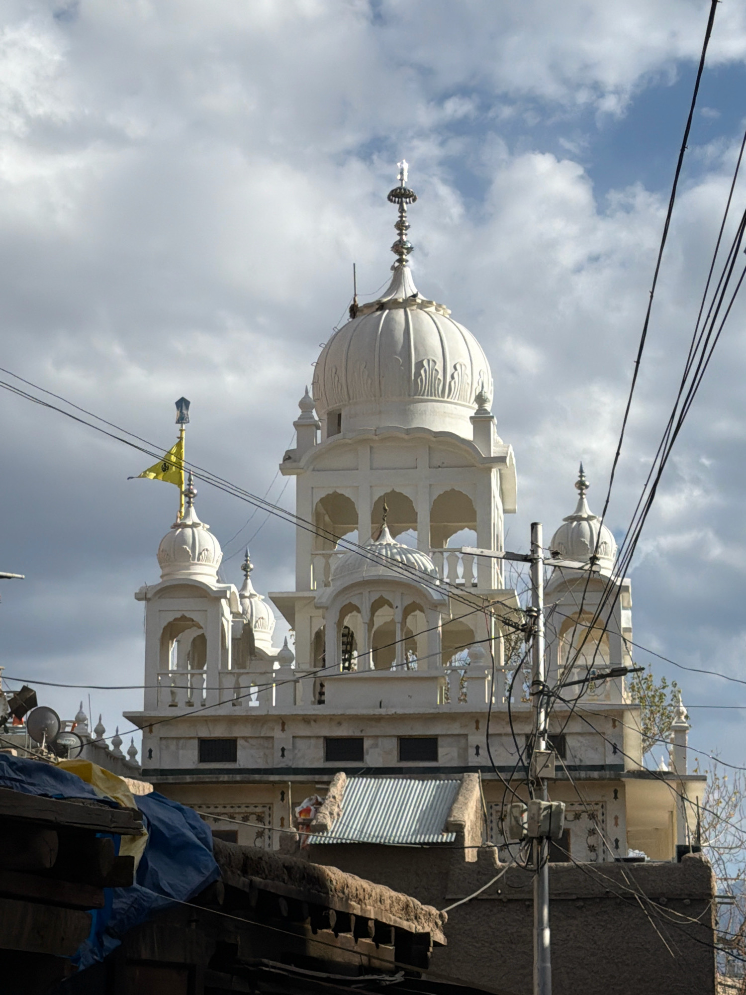 A white, multi-domed Sikh temple, identified as the Gurudwara Pathar Sahib, stands prominently against a cloudy, overcast sky in Leh, India. A yellow flag with a Sikh symbol flutters from a spire on the left side of the temple. The foreground is dominated by cluttered power lines and the rooftops of lower, less ornate buildings, suggesting an urban environment. The buildings in the foreground appear to be constructed with stone and covered with tarps and corrugated metal, indicating a more rustic or residential area. The weather appears to be fair but with significant cloud cover.