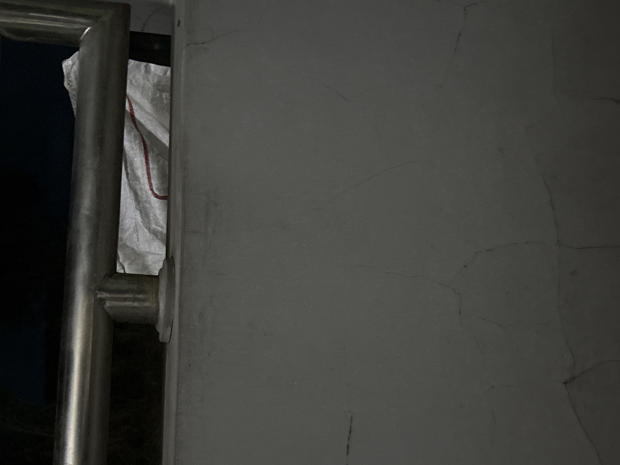 The image shows a close-up view of a gray concrete wall with visible cracks. A portion of a silver railing is visible in the upper section of the image, and behind the railing, a white bag with a red stripe is partially visible. The background is dark and out of focus. The scene is an outdoor environment, likely a balcony or an elevated walkway, located in the city of Gempol, Indonesia.