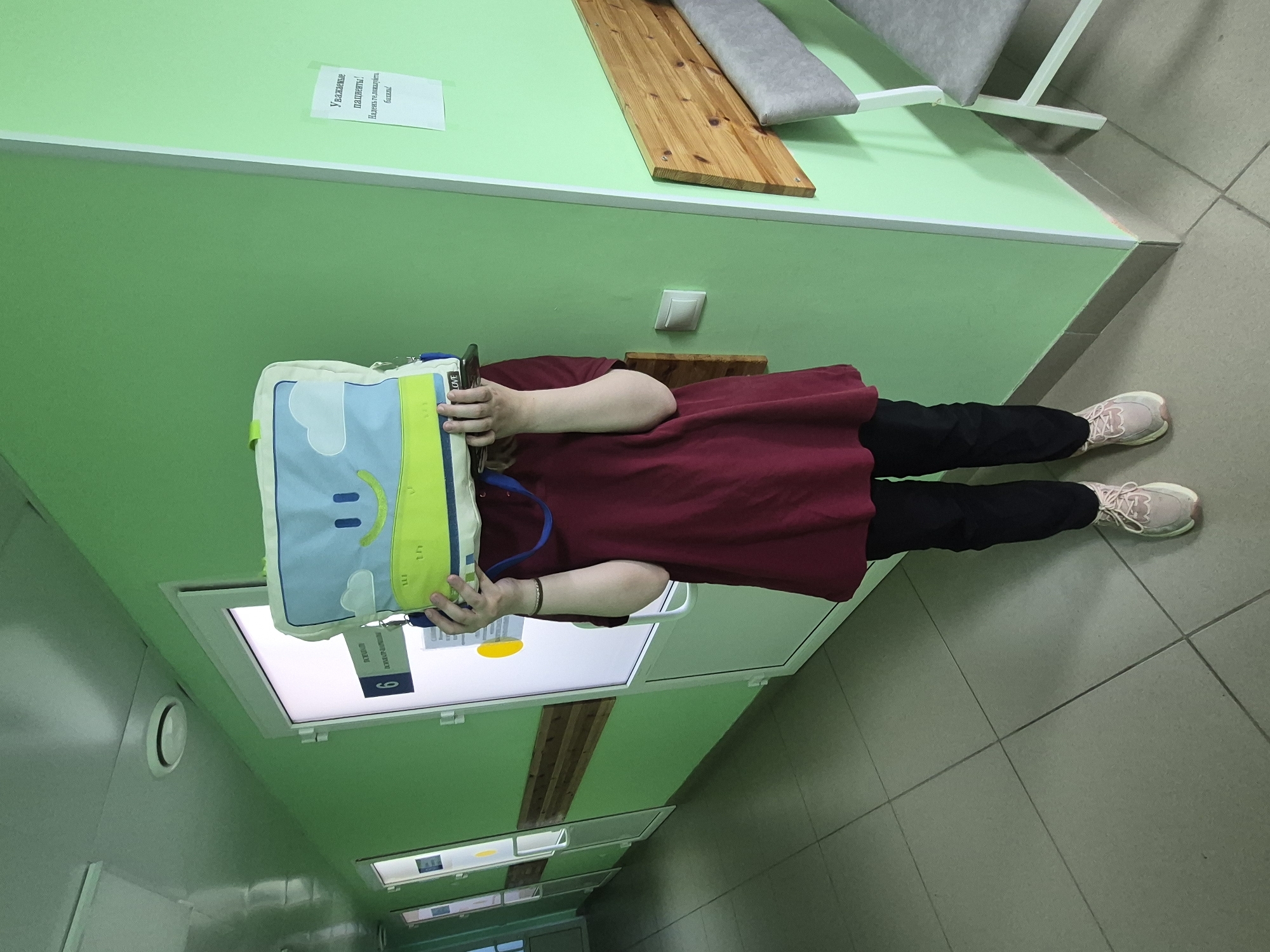 A person wearing a maroon shirt and black pants stands in a hallway with green walls and a tiled floor. They are holding a blue and green bag in front of their face. Above them is a light fixture with the number "6" visible. On the wall, there's a sign with Russian text. Wooden benches are attached to the wall.