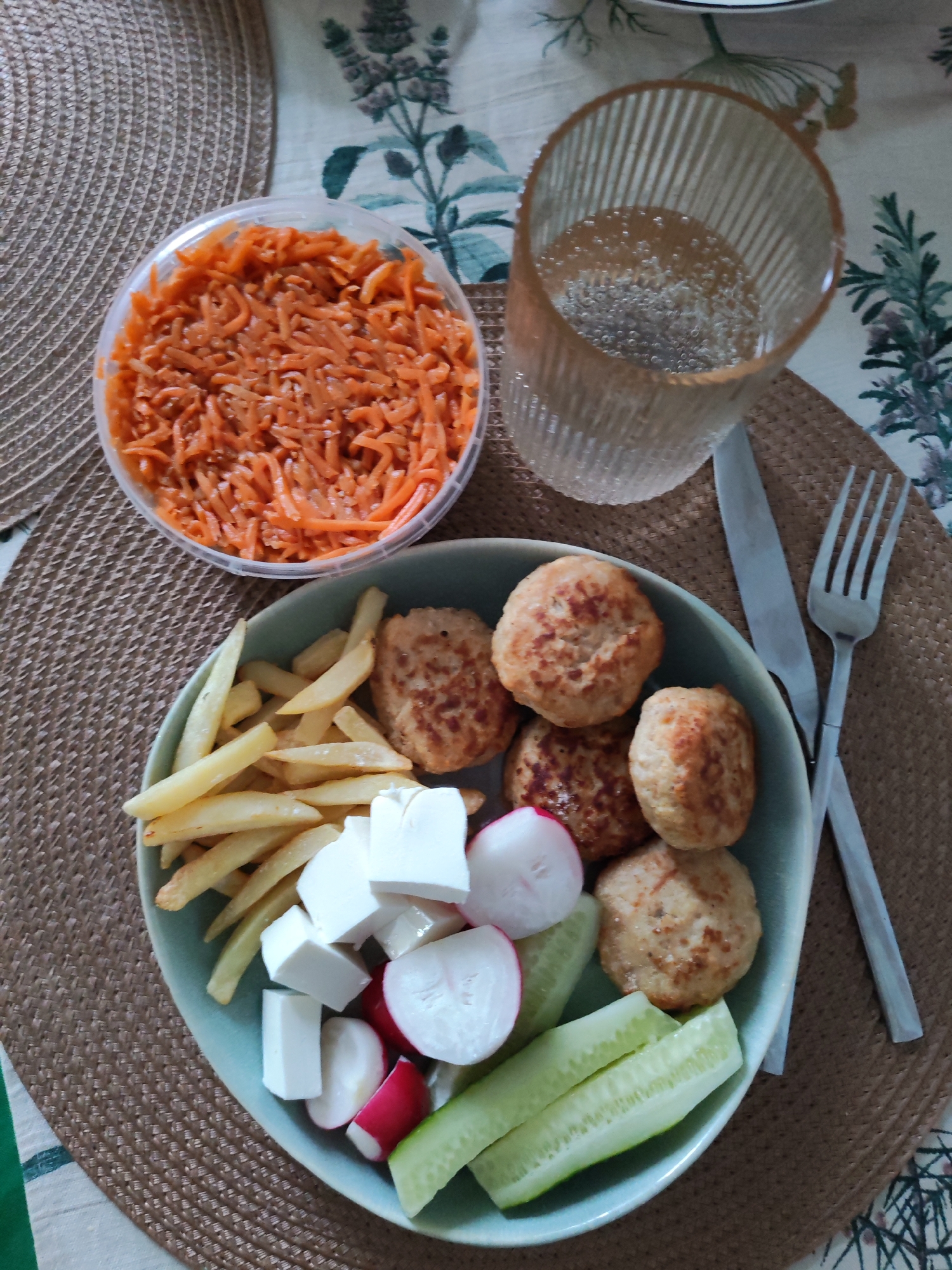 The image shows a meal on a woven placemat, with a table partially visible underneath, covered by a tablecloth with a floral pattern. The meal consists of a light blue plate filled with several components: French fries, white cheese cubes, sliced radishes, cucumber sticks, and small, round fried patties. A clear plastic container holds shredded orange carrots. A tall, clear glass filled with a fizzy liquid sits to the right of the plate. A fork and knife are placed on the placemat next to the plate. There is no information given in the picture to indicate this image being in Izhevsk, Russia.