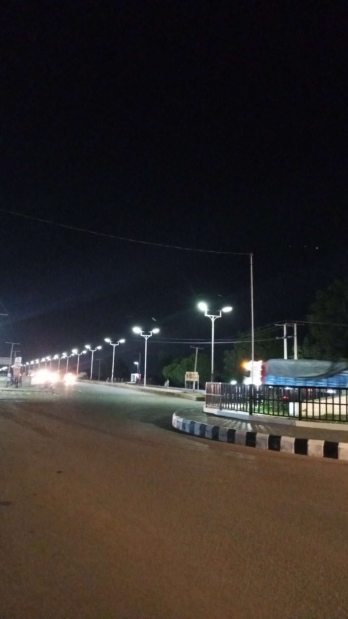 Here is a detailed description of the image:
**Setting:** An urban street scene at night. The location appears to be Unknown, Unknown.
**Environment:** The street is paved and illuminated by regularly spaced streetlights. The night sky is dark and mostly clear. Some trees are visible in the background. There is a slight curve in the road.
**Objects:**
* **Streetlights:** Tall, white poles with twin lamps are positioned along the road.
* **Road:** The road is smooth and asphalt-like.
* **Curb:** A raised curb borders the side of the road, marked with alternating black and white sections.
* **Fence:** A black metal fence runs along the side of the road.
* **Truck:** A truck is located in the distance.
**Action/Interaction:** The road appears to be mostly empty except for the truck in the distance.
**Overall Impression:** The image gives a sense of stillness and quietness due to the late hour and lack of activity on the road.