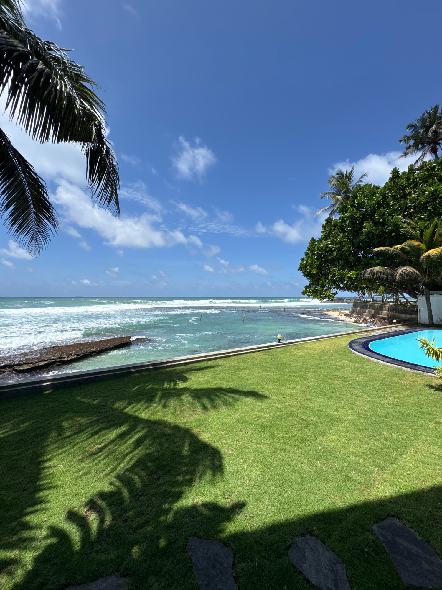 A wide shot captures a tranquil beachside scene in Goviyapana, Sri Lanka, under a bright, blue sky dotted with fluffy white clouds. The foreground is dominated by a meticulously manicured, vibrant green lawn, casting dramatic shadows from unseen palm trees. To the right, a crescent-shaped swimming pool with brilliant turquoise water is partially visible, bordered by dark blue tiling.
Beyond the lawn, a stone retaining wall separates the property from the ocean. The ocean itself is a striking aquamarine, with white-capped waves rolling towards the shore. A rocky outcropping extends into the water from the left side of the frame, creating a natural breakwater. In the distance, a few vertical poles, likely stilt fishing posts, are visible in the shallows. A single, solitary figure, a person wearing light-colored clothing, stands near the water's edge on the right side of the frame, adding a sense of scale. Lush green foliage, including a large, dense tree on the right, frames the beach and the ocean. The overall impression is one of tropical serenity and luxurious resort living.