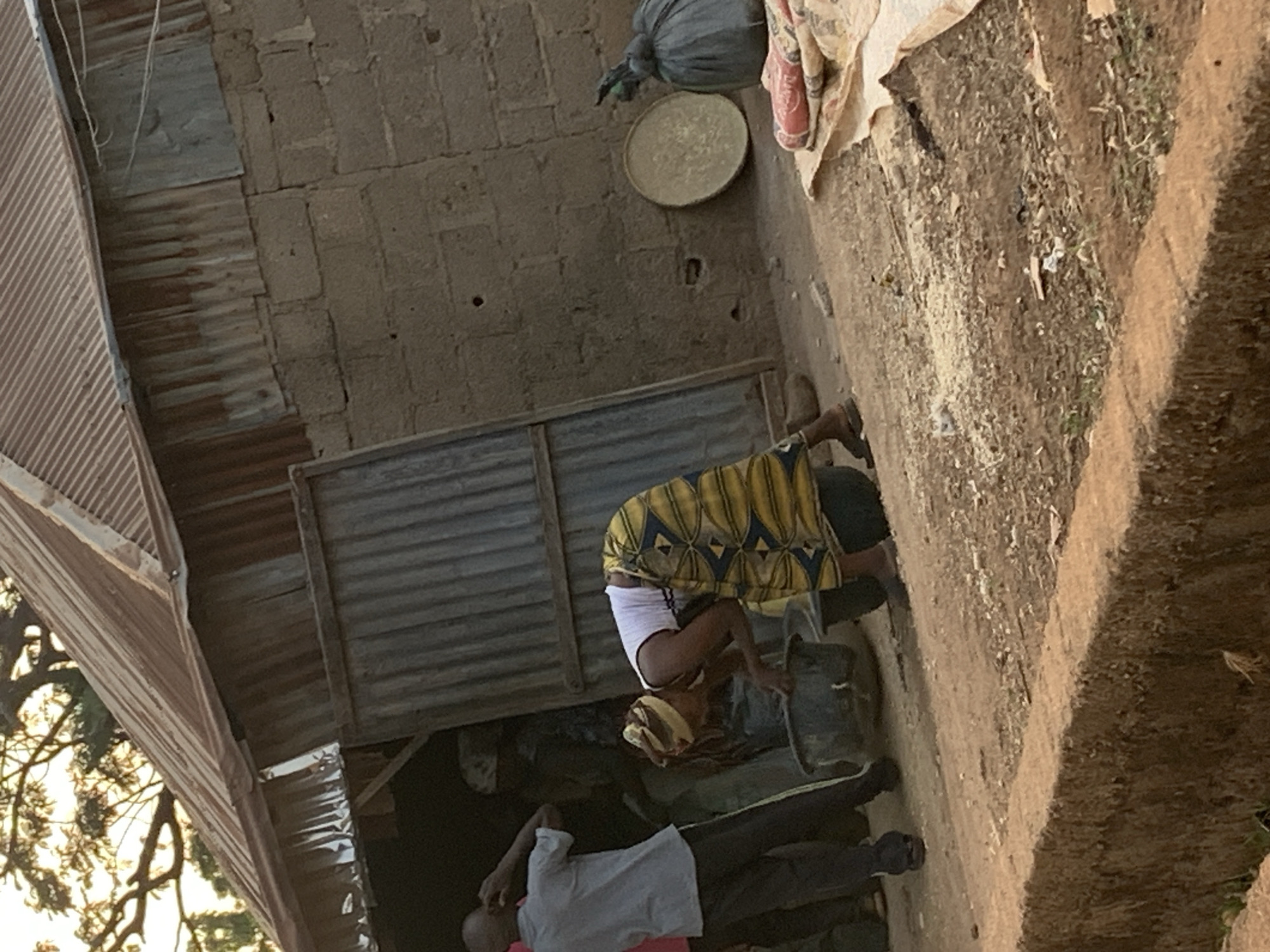 In Agban, Nigeria, a woman in a yellow and blue patterned top and white shorts is bent over a bucket outdoors. A man in a light-colored shirt and dark pants is lying on the ground nearby. A building with a corrugated metal roof and walls is in the background. A large round tray hangs on the wall, and a bag is suspended above it. The ground is dirt and vegetation.