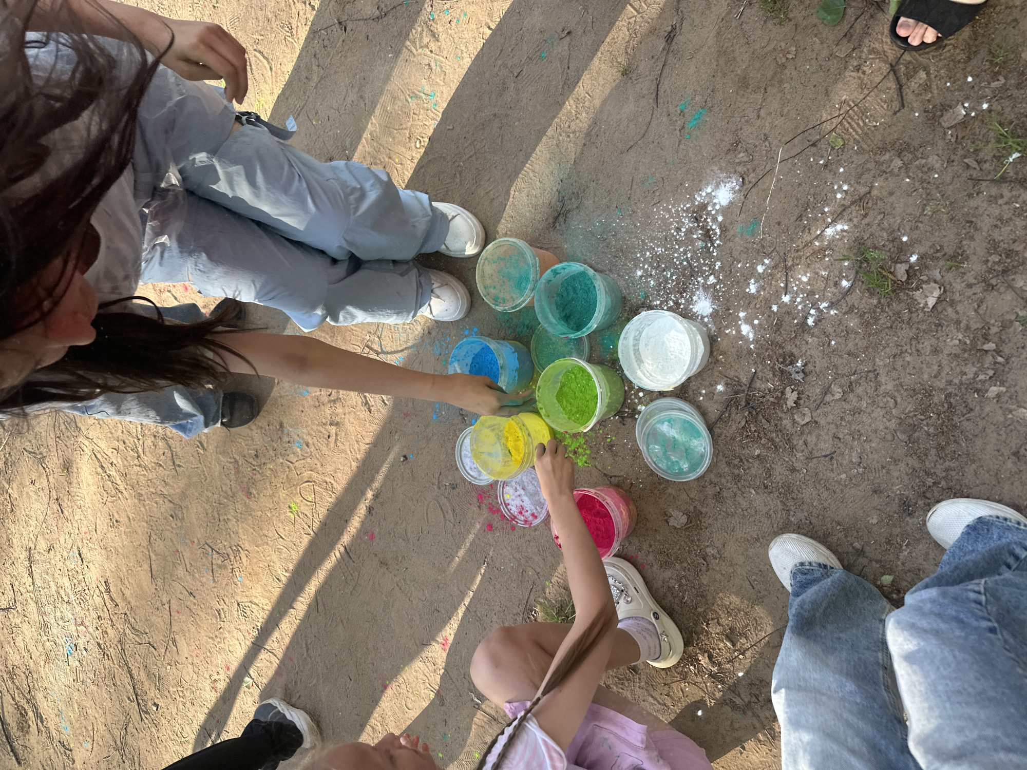 A top-down shot displays several people surrounding a collection of colorful powder containers, likely engaged in an outdoor activity. The ground is dirt, with scattered white powder, indicating that some powder has already been used.
Containers filled with blue, green, yellow, and pink powder are arranged in a circular or semi-circular pattern. Several pairs of legs and feet encircle the containers. One person is reaching into a container of yellow powder.
The people are wearing casual clothing, including light-colored pants, jeans, and sneakers. The lighting suggests that the picture was taken on a sunny day. The location is an outdoor setting, possibly a park or open space with dirt ground. The exact city and location is unknown.