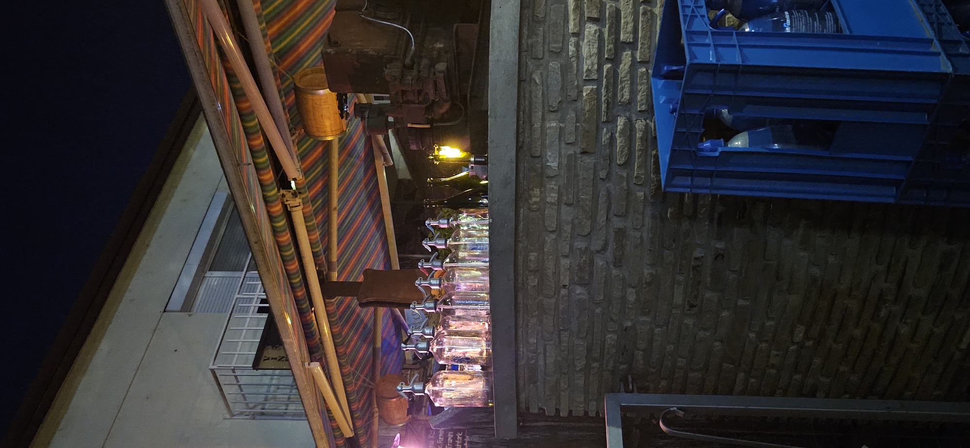 The image depicts an outdoor scene at night, likely a bar or restaurant area in Hévíz, Hungary. In the foreground, a blue plastic crate is visible, containing several large plastic bottles, some of which appear to be water bottles based on labels. Behind the crate, a wall constructed of irregular, light-colored bricks forms the base of a bar or counter. Resting on this counter are numerous glass seltzer bottles with metal spouts, arranged in a row. To the right of the seltzer bottles are two dark glass bottles and a small, lit yellow lamp. Above the counter, a striped awning in shades of red, yellow, green, and blue is supported by metal poles. In the background, a building with a light-colored facade and a window with horizontal blinds is partially visible. The sky above is dark, indicating nighttime. There are no people clearly visible in the image.
