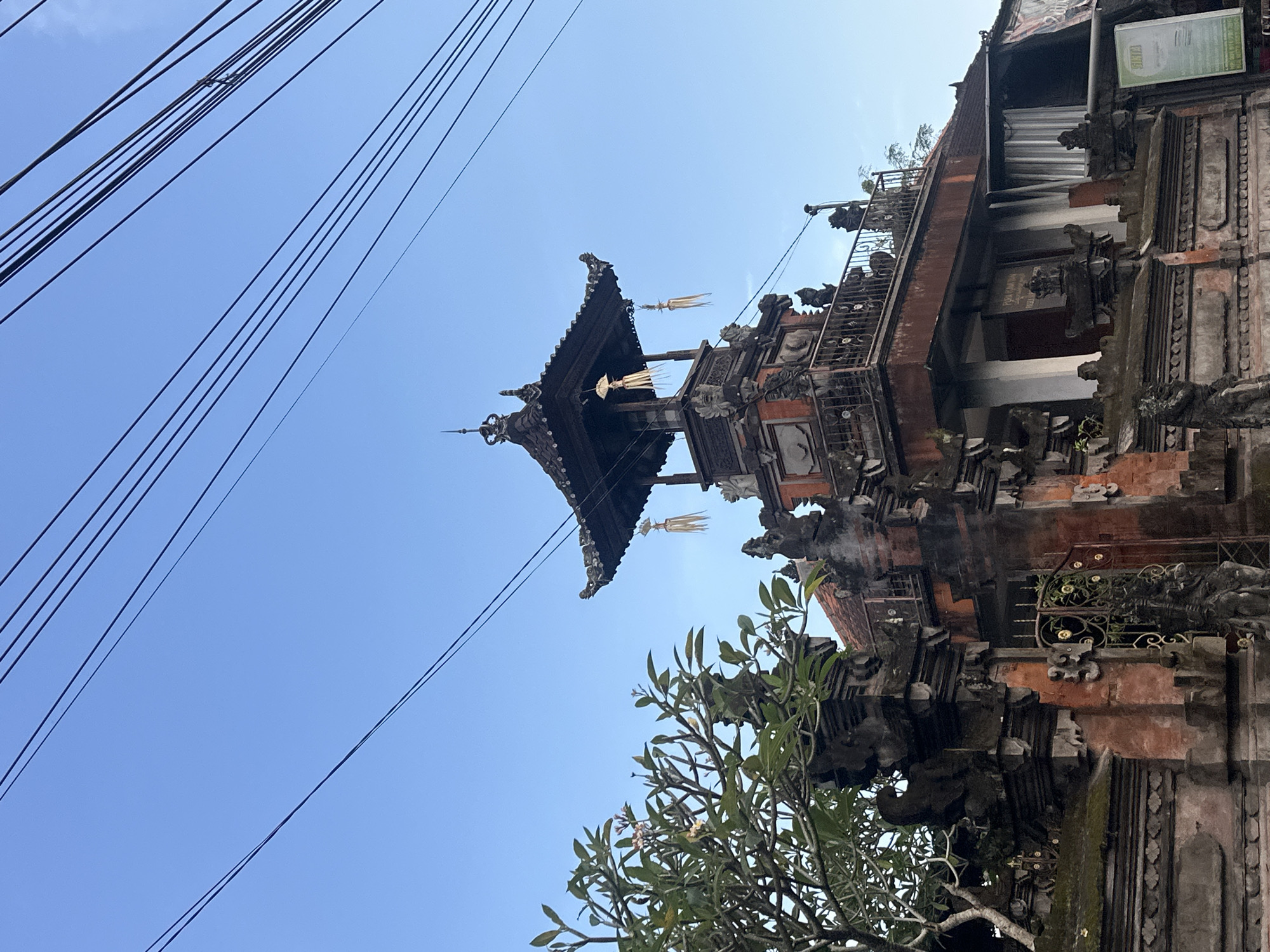 A low-angle shot depicts Balinese architecture against a bright, clear blue sky. The structure is heavily ornamented with stone carvings and weathered brickwork. Tall, decorative poles with hanging ornaments are affixed to the roof. Electrical wires run across the upper-left quadrant of the frame. Part of a tree with sparse green leaves is visible in the bottom-right corner. The location is identified as Unknown, Indonesia.
