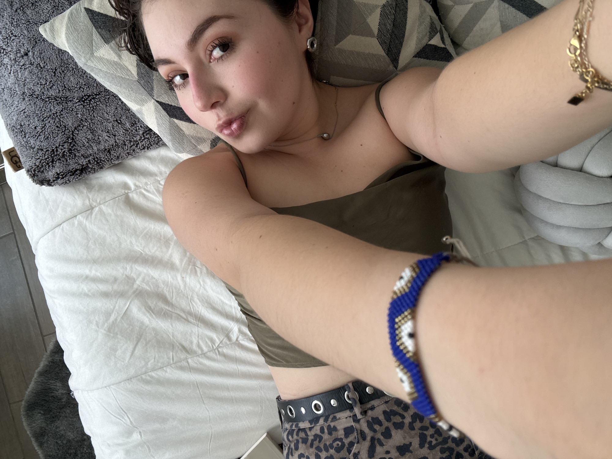 A woman is lying on a bed, taking a selfie. She has light skin, short dark hair, and is wearing a green tank top, leopard-print pants with a black belt, and jewelry, including a bracelet, earrings, and necklace. The bed has white sheets and multiple pillows, including gray textured pillows and a geometric-patterned pillow. The environment appears to be an indoor residential setting. The location is not identifiable as Mexico City.