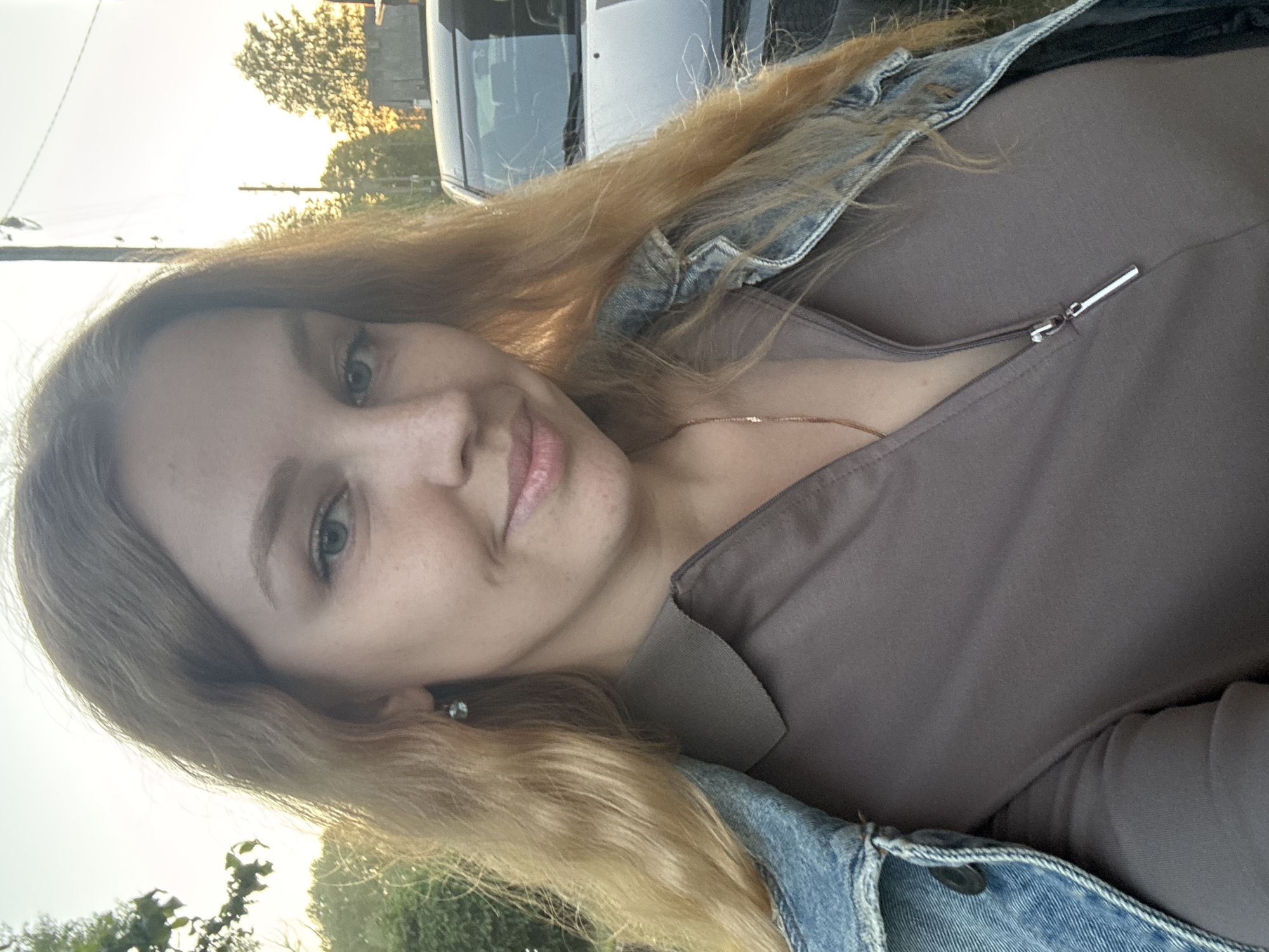 The image is a close-up, eye-level selfie of a woman taken outdoors in Rozhdestveno, Russia, under warm, natural light, likely from a setting sun. The woman has long, wavy, reddish-blonde hair parted slightly off-center and light-toned skin with visible freckles across her nose and cheeks. She has blue eyes and a faint, closed-mouth smile. She wears small, light blue stud earrings and a thin gold-colored chain necklace. Her attire consists of a brown or taupe collared shirt with a silver zipper pull, partially unzipped, and a blue denim jacket draped over her left shoulder. The background features green foliage on the left, illuminated by the warm light, and a bright, warm-toned sky. To her right, a dark-colored vehicle, possibly an SUV or truck, is partially visible, along with a utility pole and additional trees further in the distance.