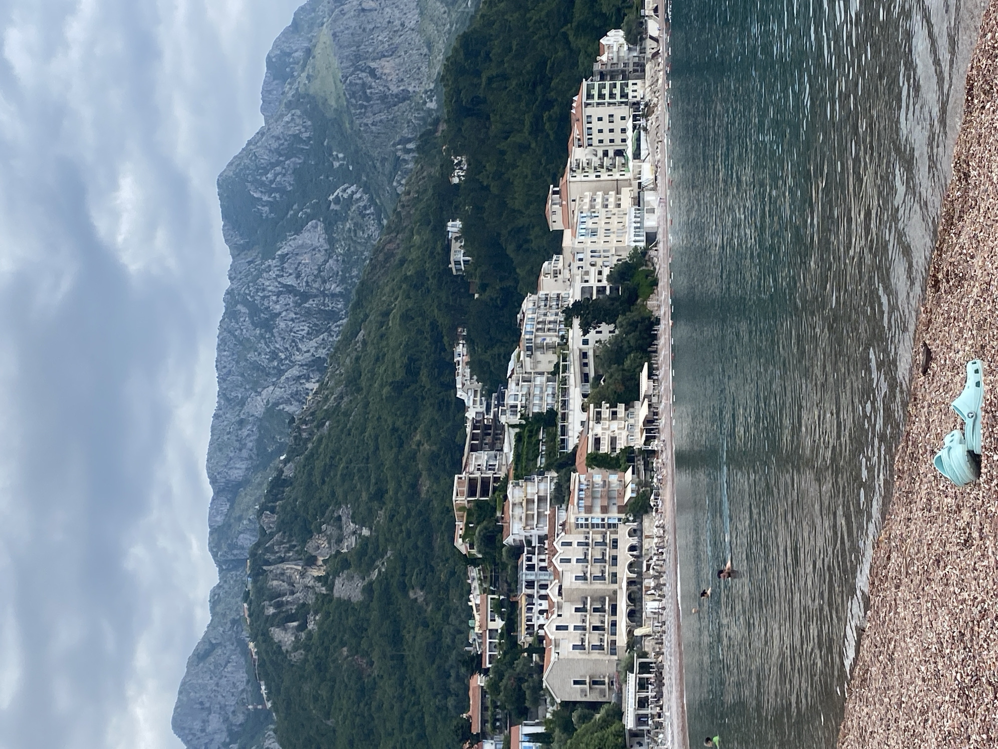 The image depicts Sveti Stefan, Montenegro. A large, gray, rocky mountain rises to the left, partially shrouded in clouds. A dense, green forest covers the lower slopes of the mountain. Adjacent to the forest, a row of tall, multi-story, beige buildings stretches along the coastline. The architecture features numerous balconies and windows. A narrow beach of small brown stones lines the edge of the water. The water is dark and rippled. At least two people are partially submerged in the water. On the right edge of the image, a pair of light blue plastic shoes rests on the stony beach.