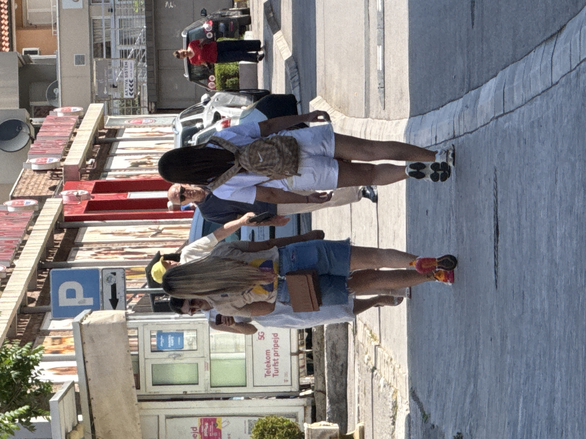 The image depicts a street scene in Budva, Montenegro, likely during daylight hours given the bright sunlight. Two women, one with blonde hair and wearing denim shorts and a t-shirt, and another with dark hair and wearing white shorts and a t-shirt with a backpack, are walking away from the camera on a paved sidewalk. The blonde-haired woman is carrying a small brown crossbody bag. Behind them, a man with a yellow cap is visible, and further back, a man with sunglasses and a dark shirt is walking. Several cars are parked or driving on the street to the right, and buildings line the left side of the street, some with signage. One sign reads "Telekom 5G Turist pripejd." A blue parking sign with a white "P" and an arrow pointing left is also visible. The environment appears to be an urban setting with commercial establishments.