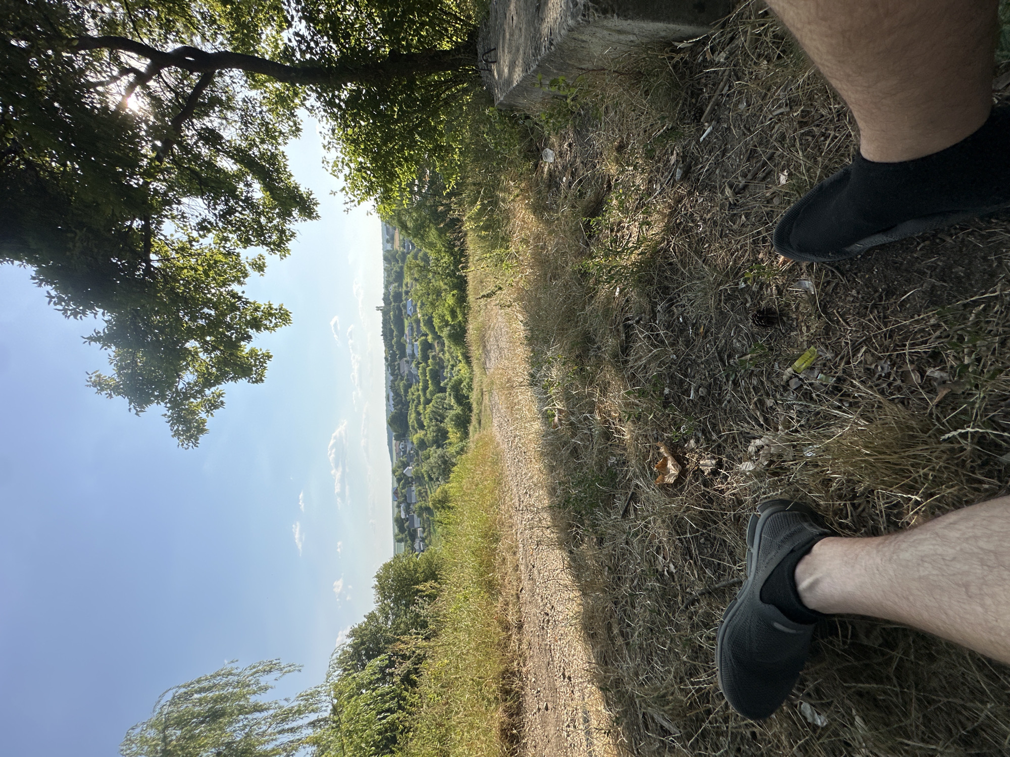 An outdoor, eye-level, full shot shows a person's legs and feet resting in the foreground, with a scenic overlook of a rural town in the background. The person is sitting on a grassy incline, with their legs extended forward and their feet clad in dark athletic shoes and socks. The ground is covered in dry grass, with a scattering of fallen leaves and pinecones. A dirt path leads up the incline to the left, and a large, weathered concrete block is visible to the right, partially covered by vegetation.
Behind the foreground, the landscape slopes down to reveal a town nestled among rolling green hills. Numerous houses with red and white roofs are visible, interspersed with trees. In the distance, a church spire and other structures hint at the town's layout. The sky above is a clear, vibrant blue with a few wispy white clouds. Sunlight filters through the branches of a large tree on the right, casting dappled shadows on the ground. The overall impression is one of a peaceful, sun-drenched afternoon overlooking a Ukrainian village. The specific location is in the vicinity of Makiv, Ukraine.