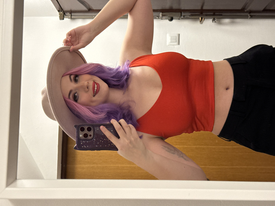 A woman poses in a mirror selfie, tilting her head and body. She wears a tan hat, a red tank top, and black pants. Her hair is a vibrant mix of pink and purple hues. She holds a phone with a purple, starry patterned case. The background features a white wall with electrical fixtures and a wooden door. The image is framed by the white edge of the mirror.
