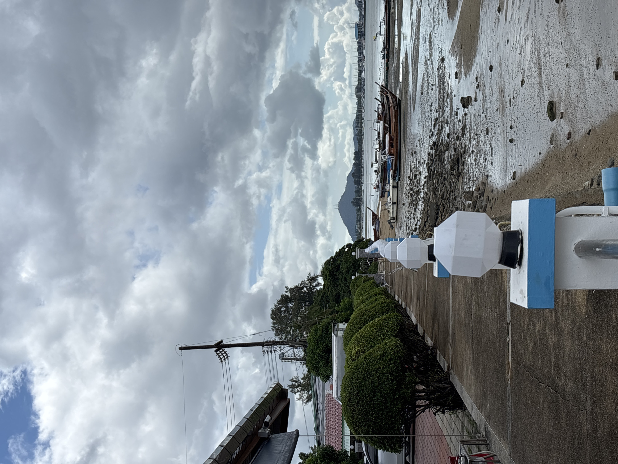 The image depicts a waterfront scene in Rawai, Thailand, under a cloudy sky. A concrete walkway runs along the left side of the frame, lined with neatly trimmed green bushes. Beyond the bushes, a building with a dark roof and a car are visible. A utility pole with numerous wires stands tall in the background.
The right side of the walkway features a series of white outdoor lights with black bases, mounted on white posts with blue trim. The walkway overlooks a muddy, rocky shoreline where several wooden longtail boats are moored. The tide appears to be out, exposing a large expanse of wet sand and exposed rocks. In the distance, more boats and structures can be seen across the water, with a hilly landscape forming the horizon. The overall atmosphere is overcast and somewhat muted.