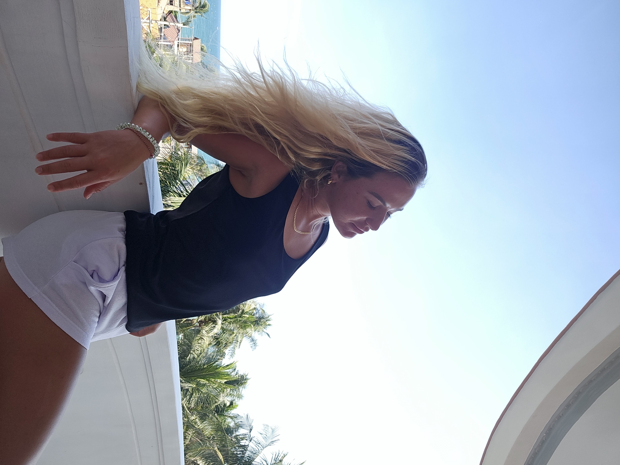 A blonde woman with her eyes closed leans on a white railing on a balcony. She is wearing a black tank top and white shorts. Her long blonde hair is blowing in the wind. In the background, there is a view of the ocean and palm trees, suggesting a tropical location. The sky is clear and blue. Based on the context of Phu Thuy Ward, Vietnam, this scene likely depicts a resort or a residential balcony overlooking the coastline.