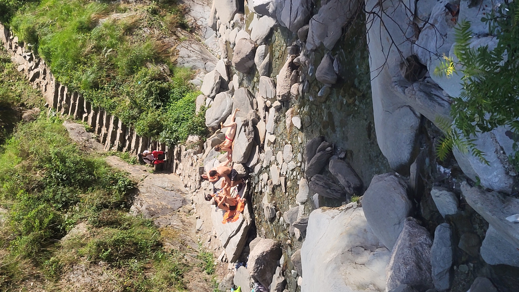 The image captures a group of people interacting within a natural setting in Bhagsu, India. The scene is dominated by a rocky, mountainous terrain, including a stream or riverbed filled with various-sized rocks and stones. Several individuals are positioned on the rocks, with some appearing to be relaxing or sunbathing. One person seems to be preparing to jump into the water.
To the left of the frame, a constructed pathway or staircase ascends the hillside, lined with vegetation. A person wearing red clothing is visible on the path. The general environment suggests a mountainous region.
