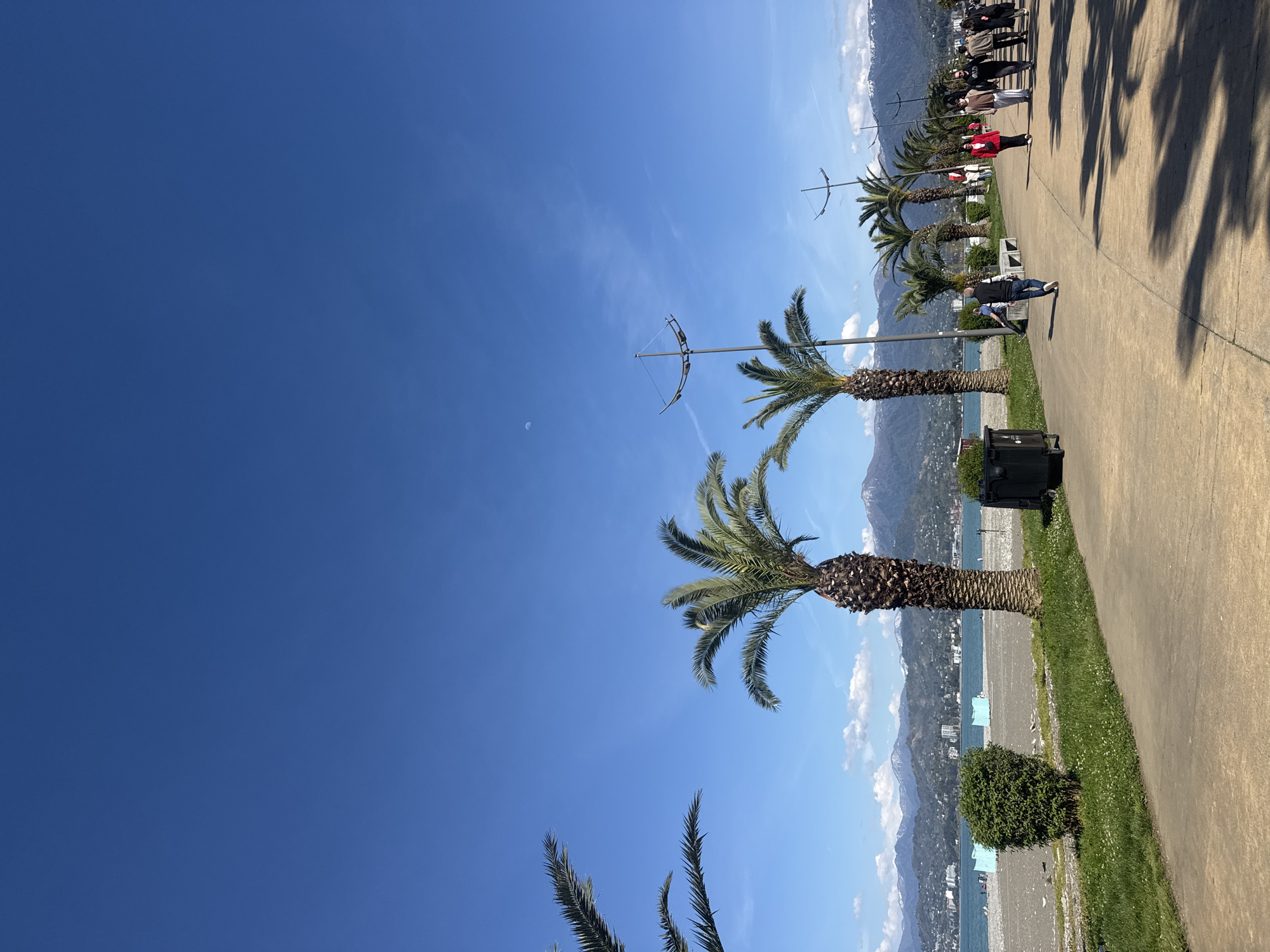 The image captures a sunny day along a coastal promenade. Palm trees line the walkway, casting shadows on the paved path. A small patch of grass separates the walkway from the beach below. Distant mountains rise in the background, with buildings visible along the shoreline. People can be seen walking along the promenade. The sky is a clear, bright blue. The image appears to be taken in Batumi, Georgia.